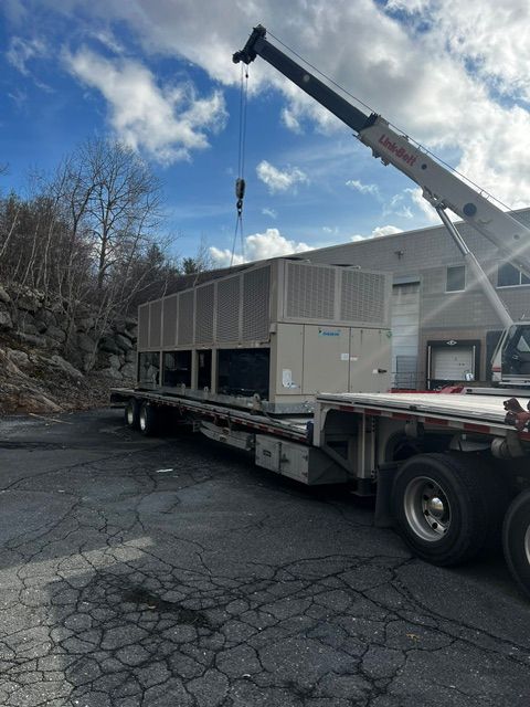Flatbed truck crane lifting a large white HVAC unit in a cracked parking lot under a partly cloudy sky