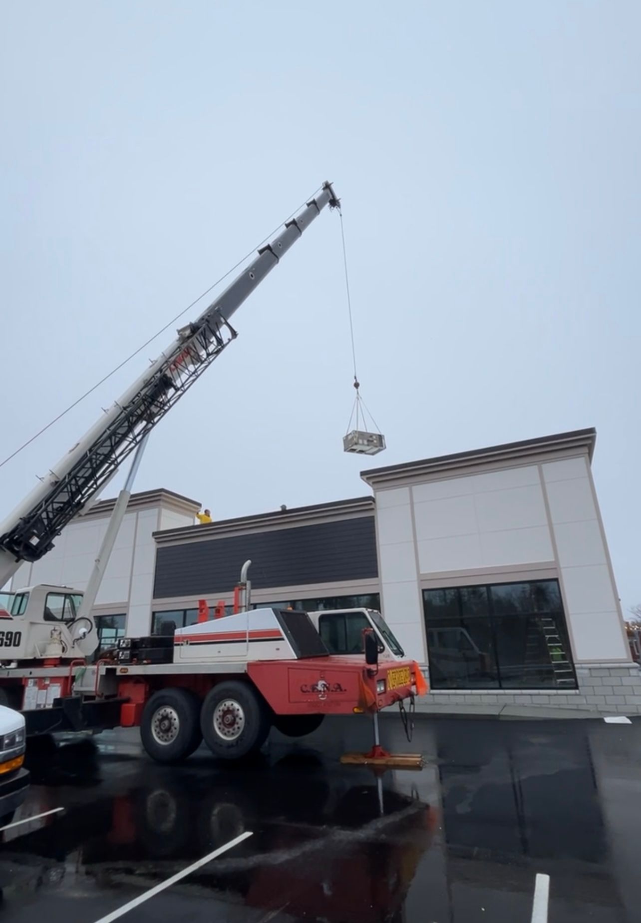 Crane lifting a small load beside a white building on a wet street.