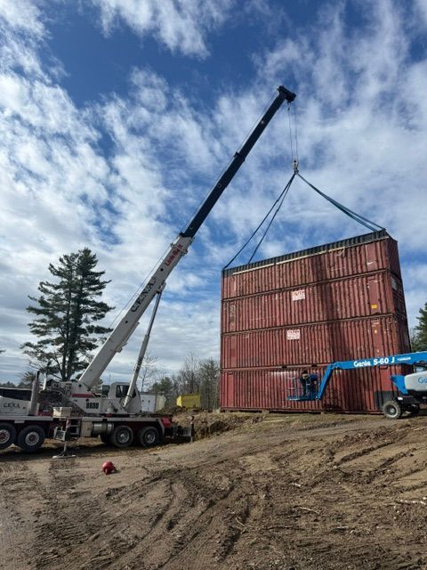 Crane lifting a red shipping container at a muddy construction site under a cloudy sky