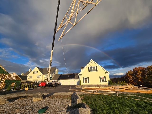 Crane lifting a roof truss at a construction site beside two yellow houses under a cloudy sky