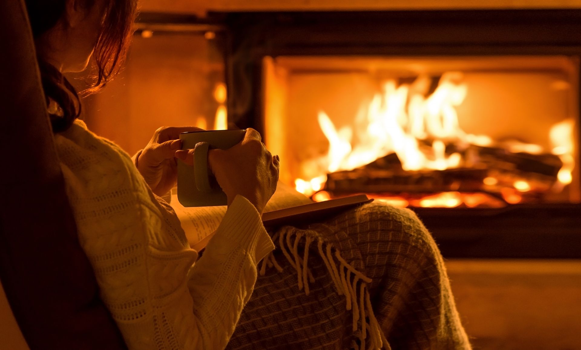 A person sits in a cozy chair by a warm fireplace, holding a mug while wrapped in a blanket.