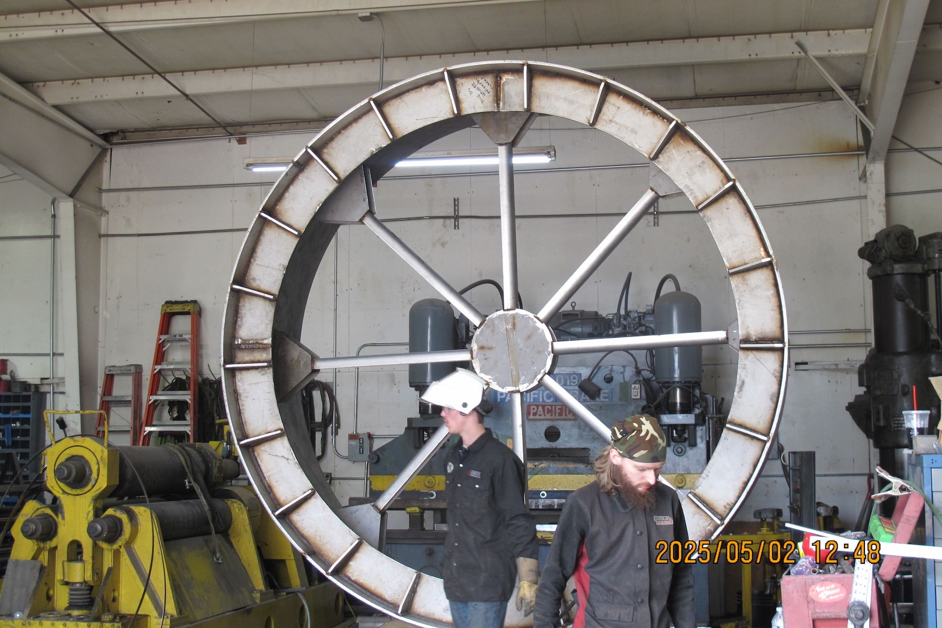 Two men are standing in front of a large metal wheel
