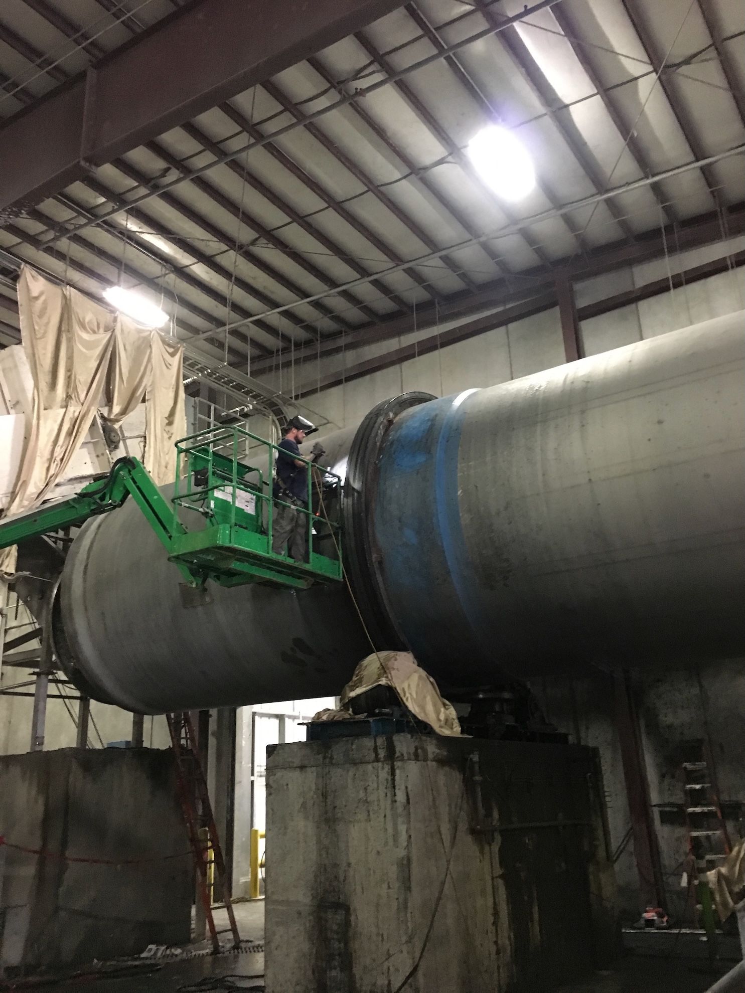 A man on a crane is working on a large pipe in a factory.