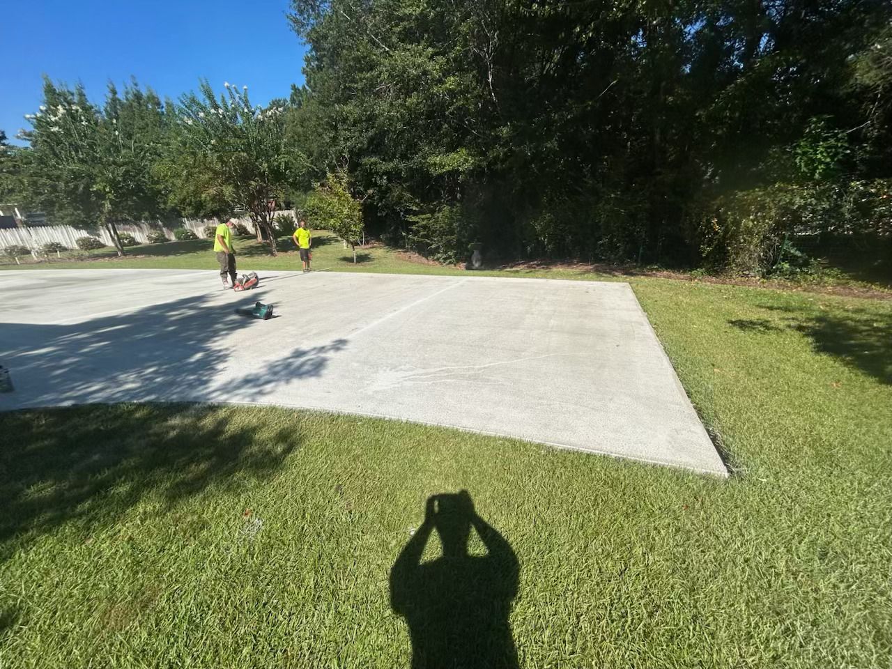 Two people stand on a concrete pad in a grassy area, trees in the background, a person's shadow in the foreground.