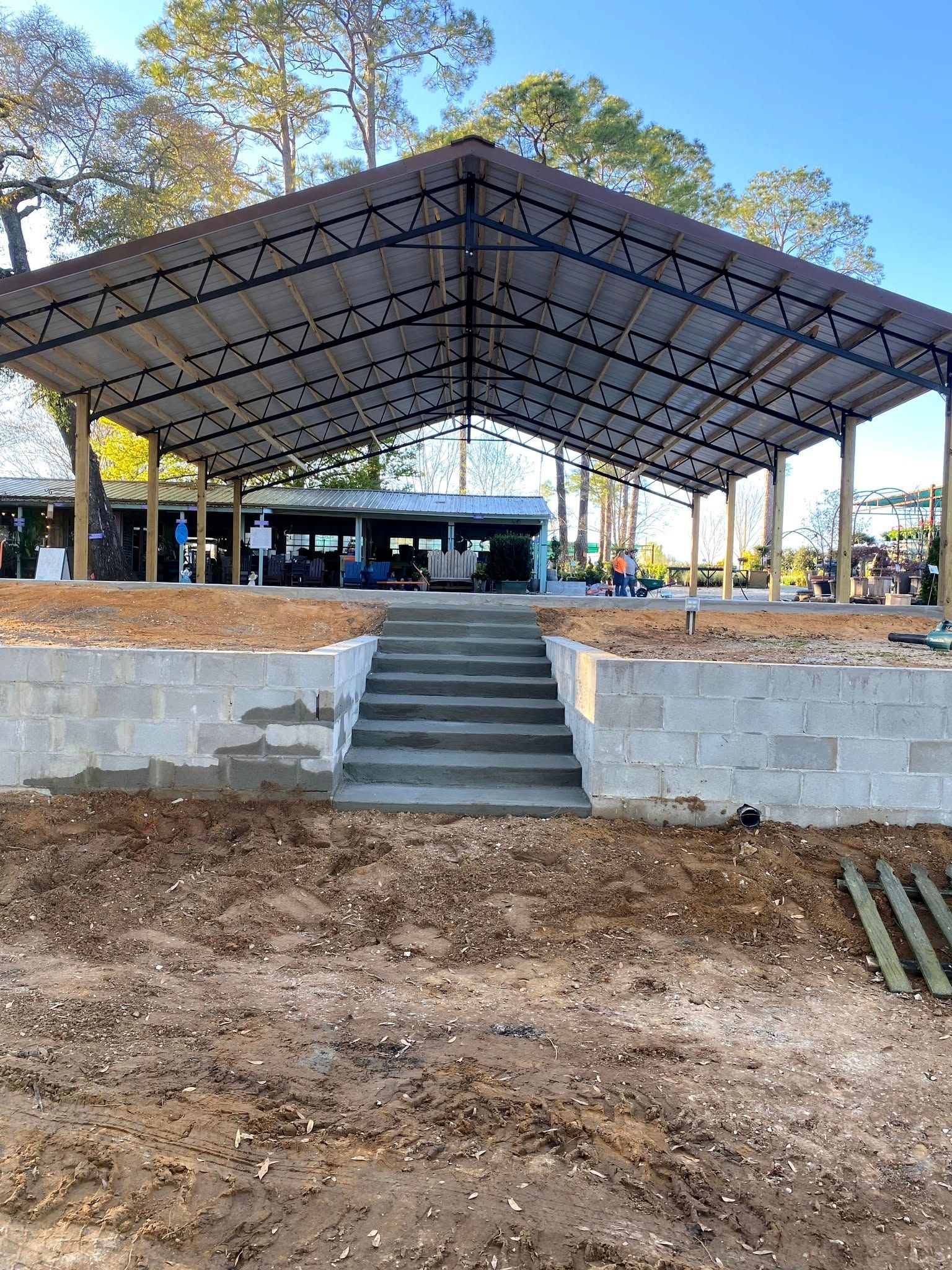 Construction of a covered pavilion with concrete steps. Dirt ground, blue sky, and trees.