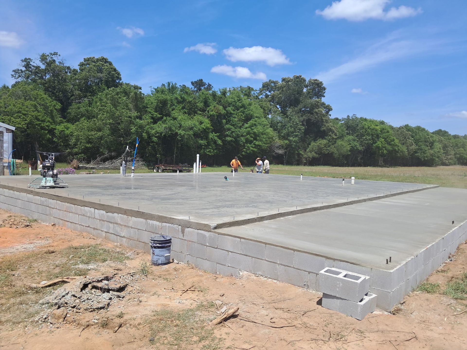 Newly poured concrete foundation with cinder block perimeter, surrounded by grass and trees under a blue sky.