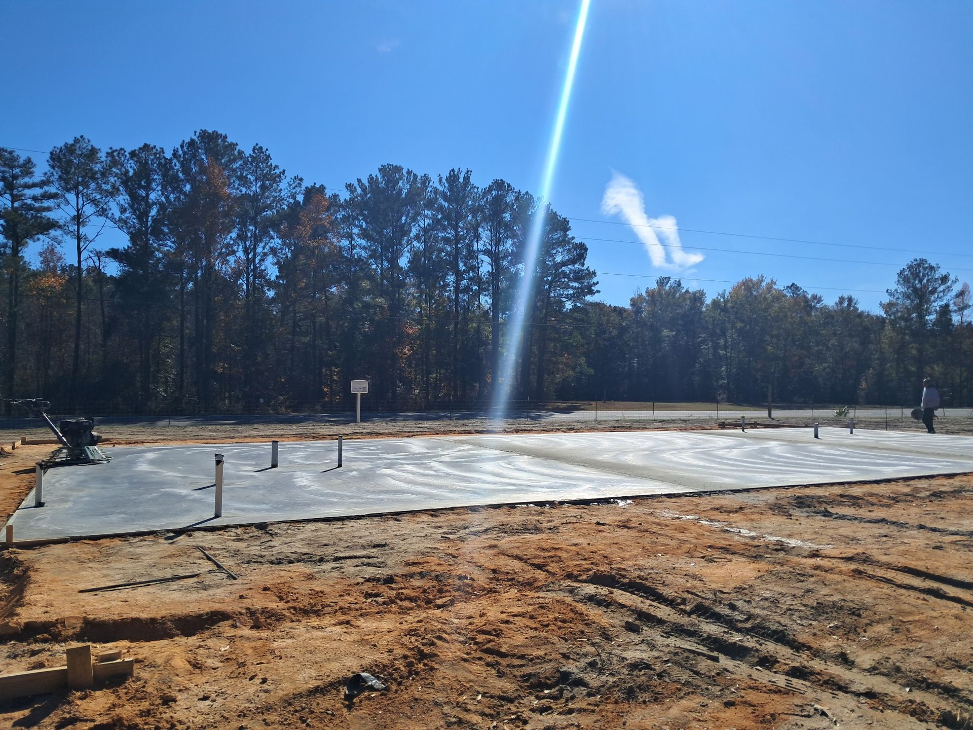 Concrete foundation with exposed pipes, prepared construction site; trees and blue sky background.