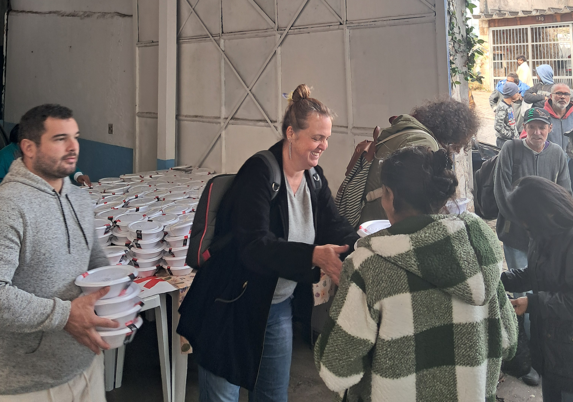 Pessoas distribuindo refeições para um grupo. Uma mulher sorri e aperta as mãos. Outros seguram recipientes de comida.