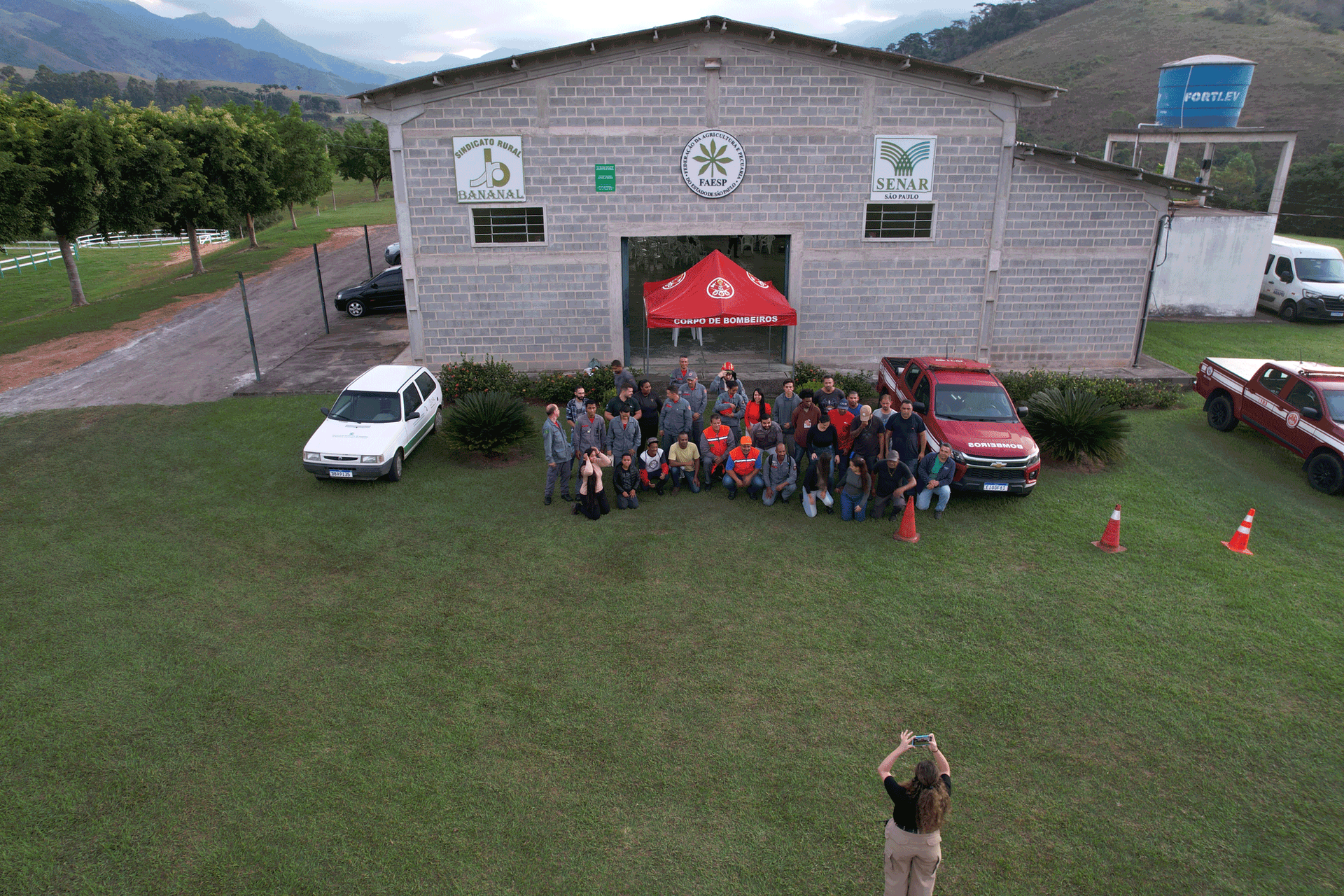 Grupo de pessoas, incluindo bombeiros, posando em frente a um prédio com uma tenda vermelha, cercado por veículos.