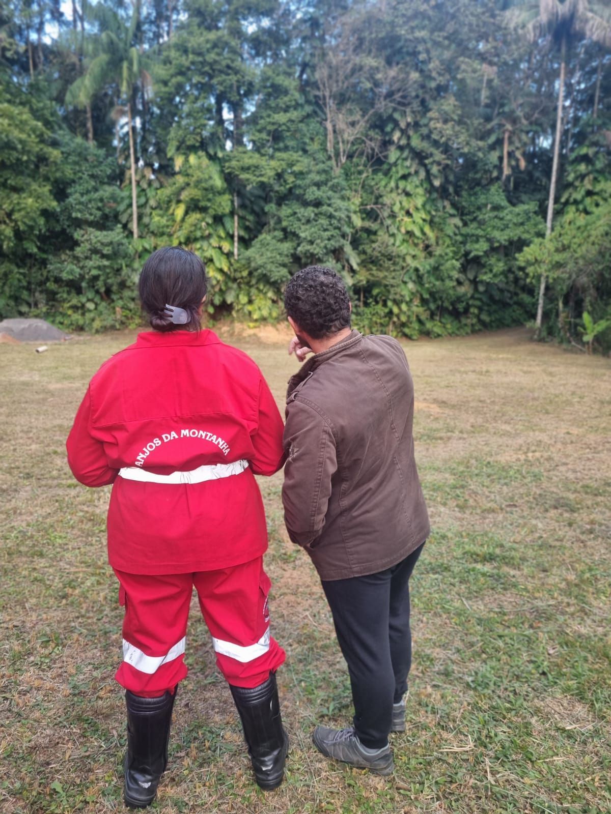 Duas pessoas olhando para as árvores; uma delas usa um macacão vermelho com detalhes brancos e botas pretas.