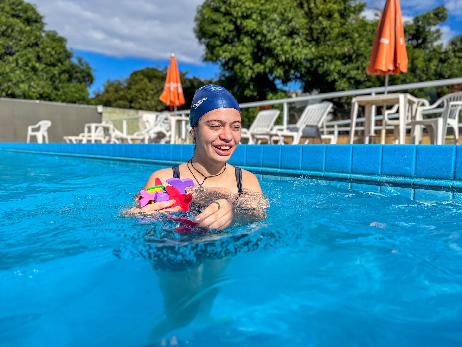 Mulher de touca de natação azul, sorrindo em uma piscina, segurando brinquedos.