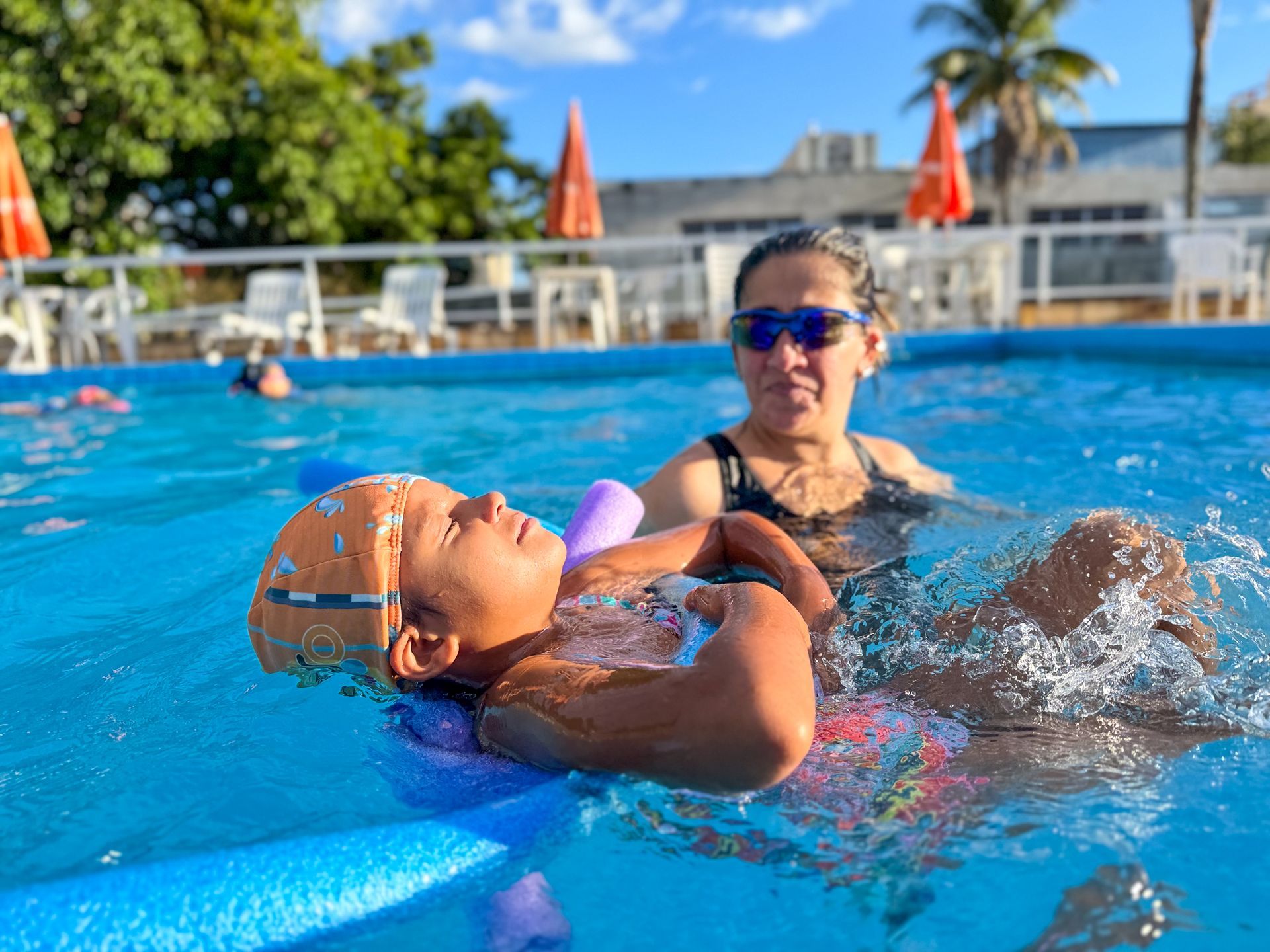 Mulher ajudando uma criança a flutuar em uma piscina azul brilhante; ambas vestindo trajes de banho sob um céu ensolarado.