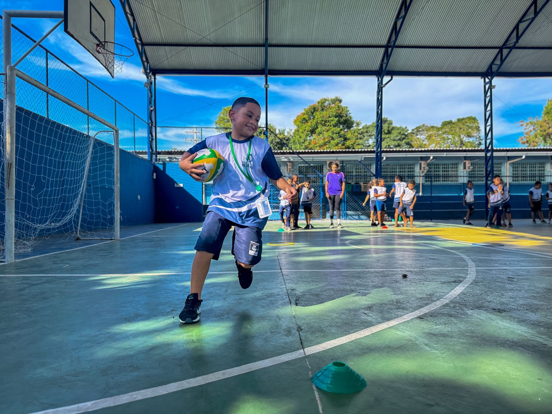 Menino corre com uma bola na quadra da escola, sorrindo. Outras crianças observam. Azul, verde e ensolarado.