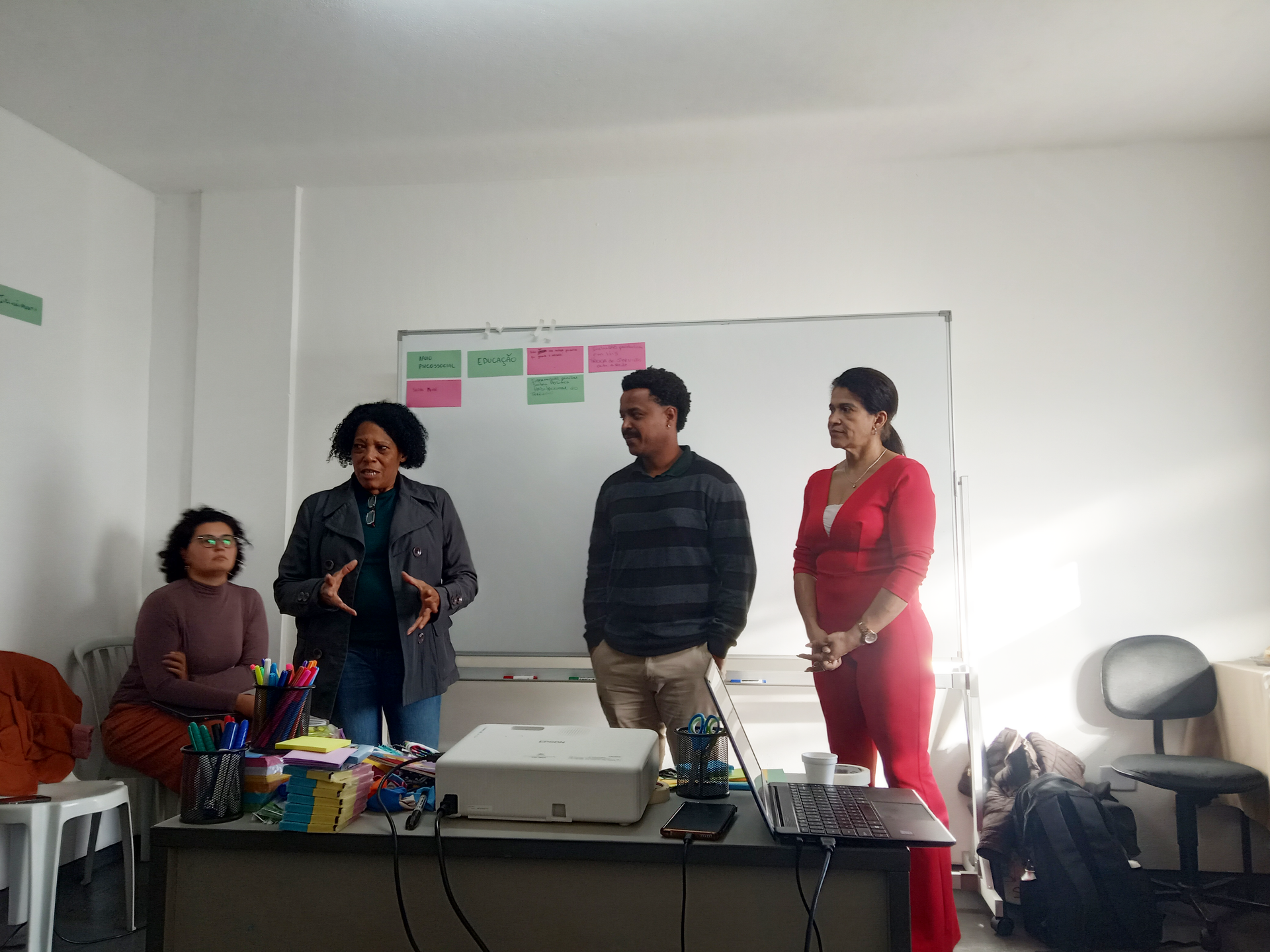 Quatro pessoas estão em frente a um quadro branco; uma mulher fala com as mãos enquanto as outras ouvem.