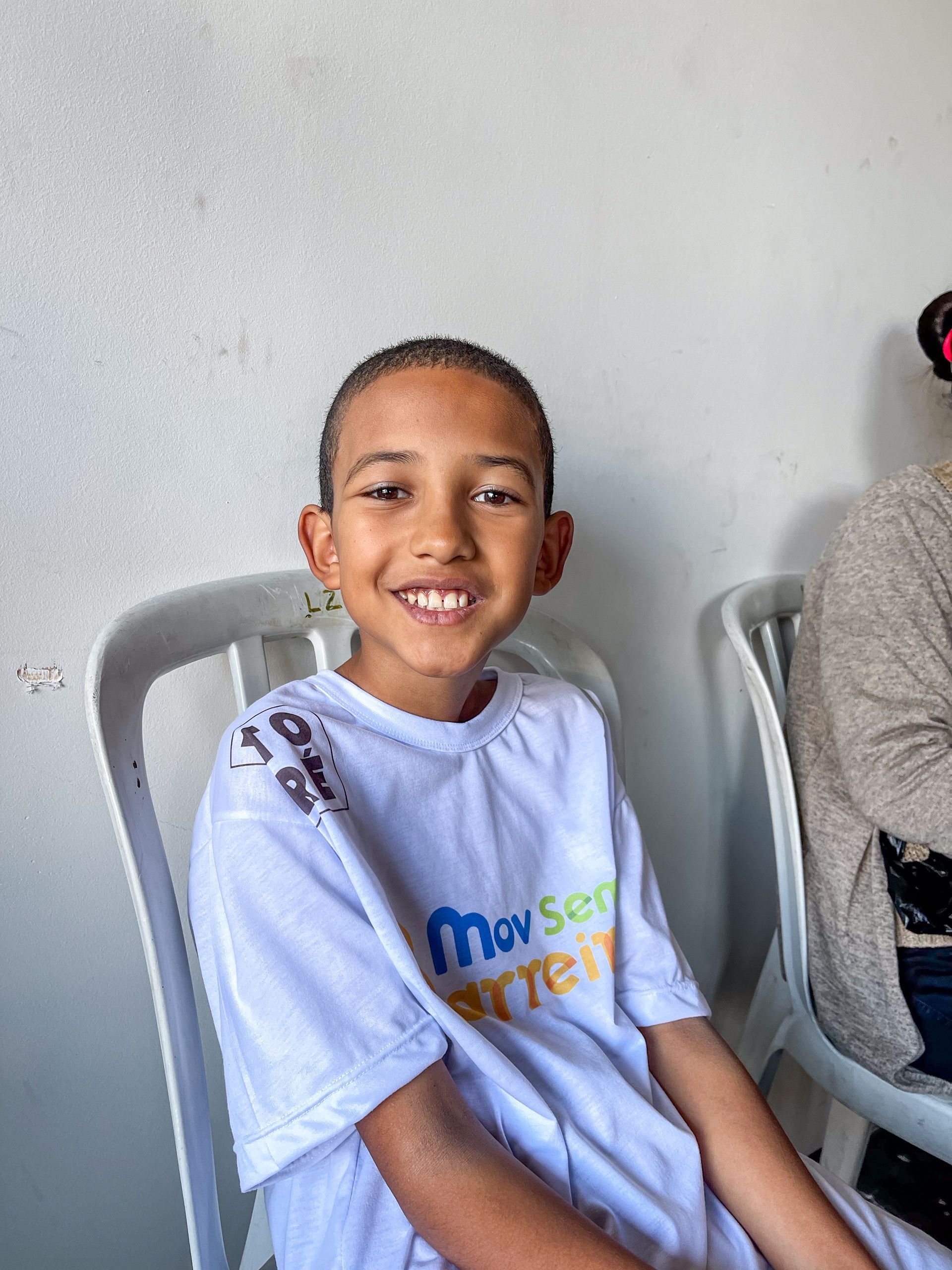 Menino sorrindo, vestindo camiseta branca, sentado em uma cadeira, encostado em uma parede branca.