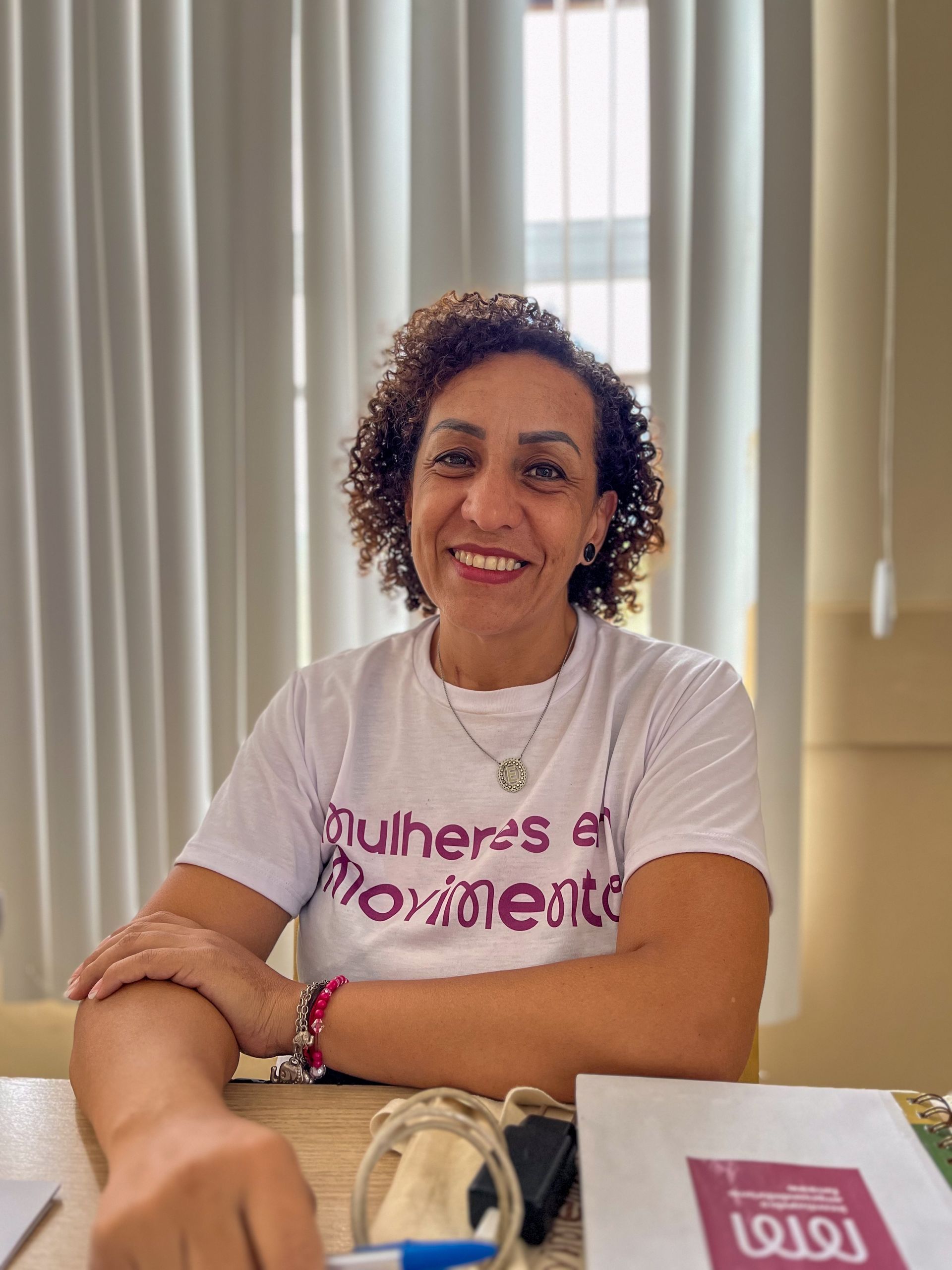 Mulher sorridente, de cabelos cacheados, vestindo uma camisa branca, com a inscrição “Mulheres em Movimento”, sentada à mesa.
