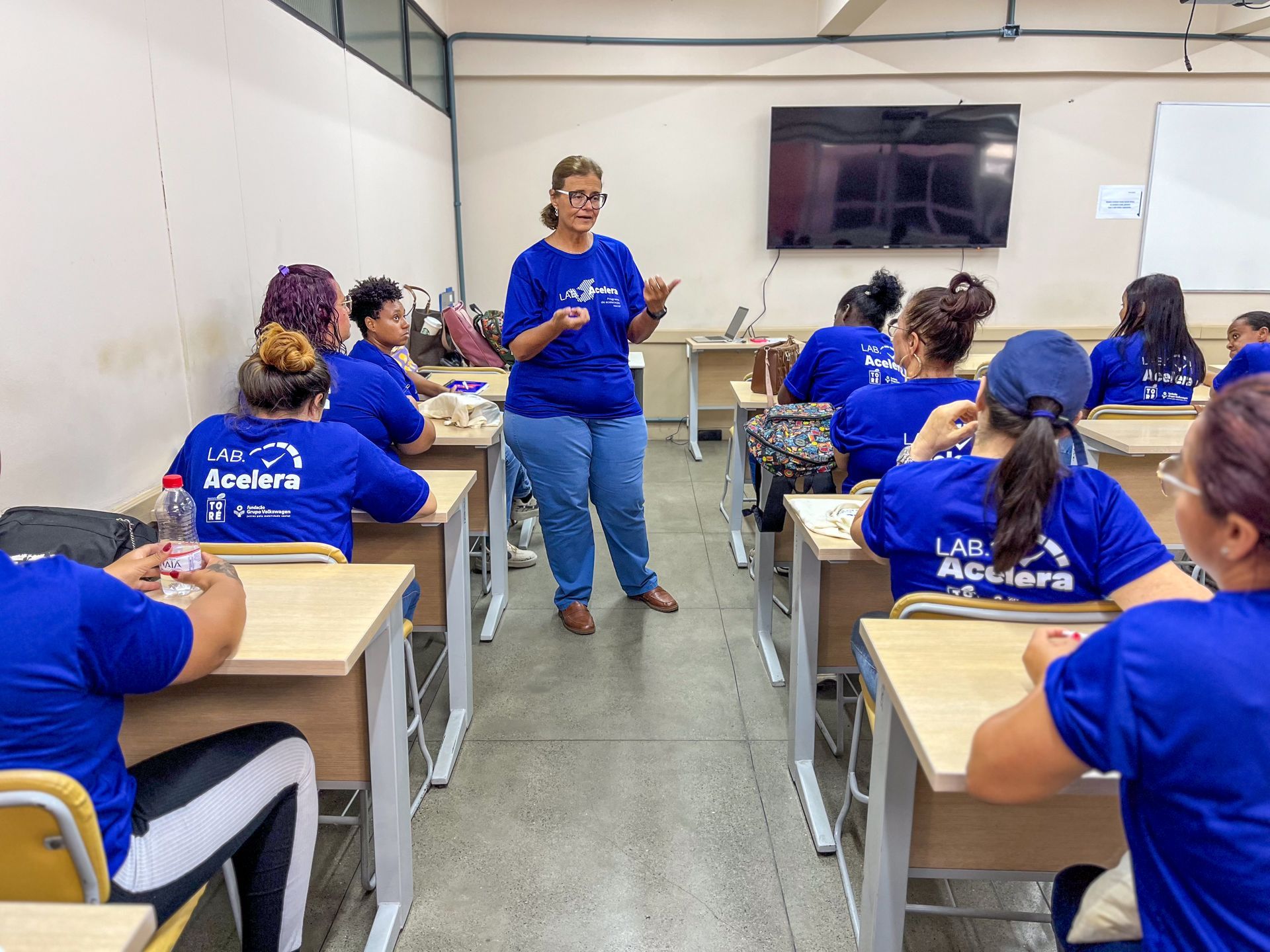 Mulher de blusa azul dá aula para alunos em uma sala de aula. Alunos usam camisas azuis e carteiras dispostas em fileiras.