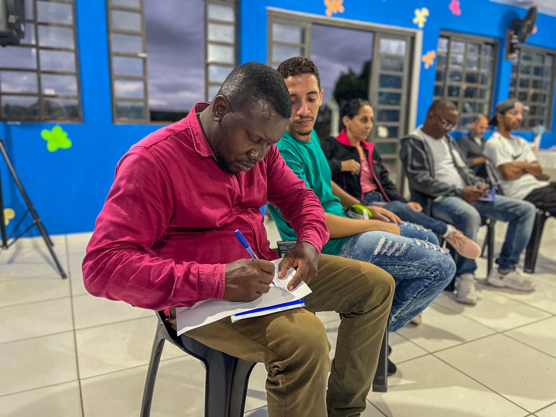 Homem de camisa rosa escrevendo, sentado com outros em uma sala de paredes azuis, provavelmente uma reunião ou workshop.