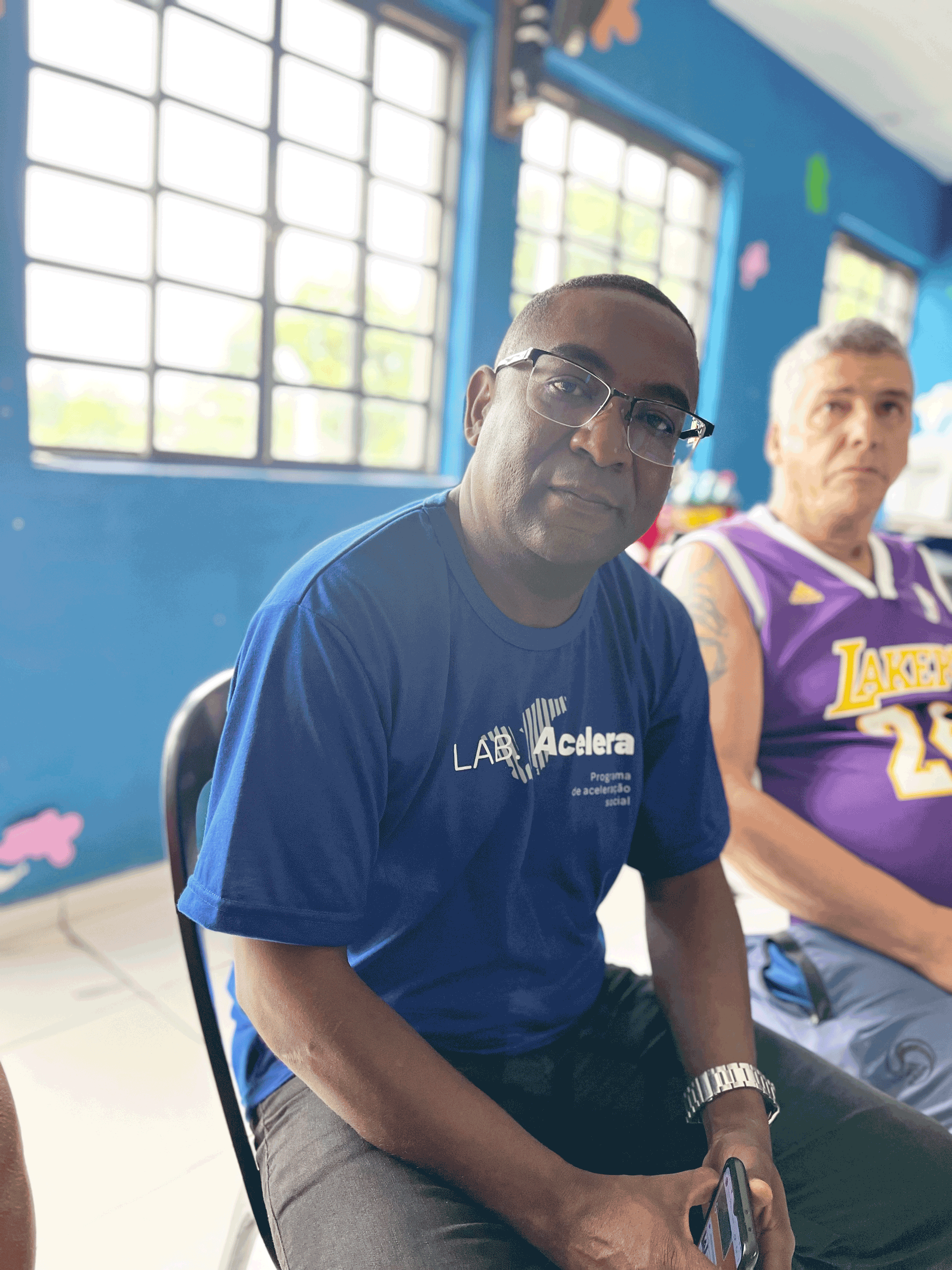 Homem de camisa azul e óculos olhando para a câmera, sentado perto de outro homem vestindo uma camisa dos Lakers.