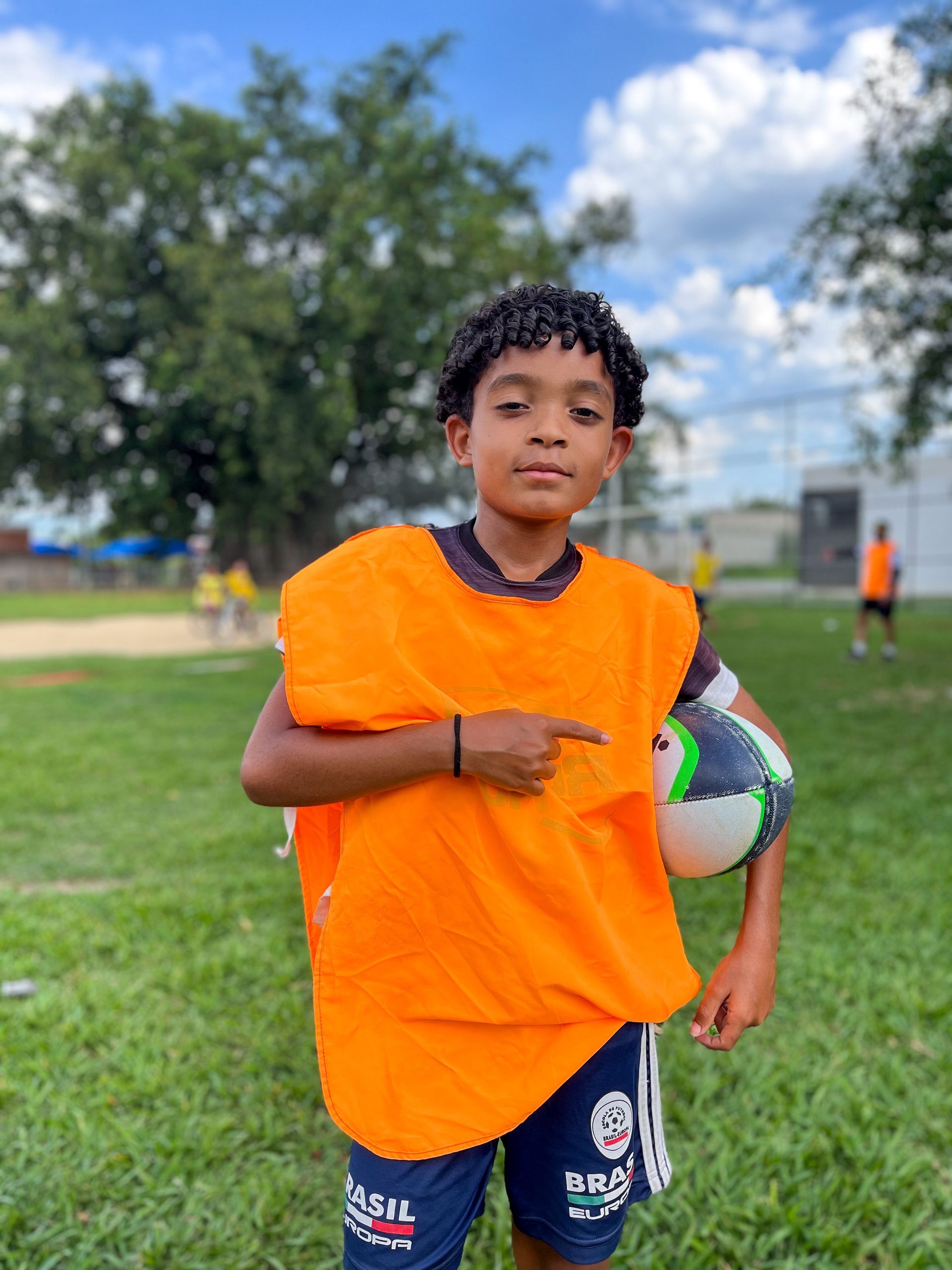 Menino de colete laranja segura bola de futebol e aponta em um campo gramado.