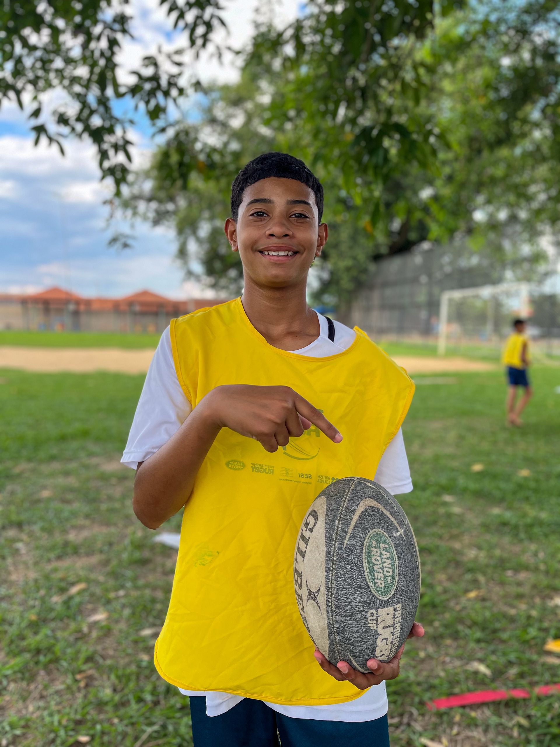 Jovem de colete amarelo apontando para uma bola de rugby em um campo gramado.