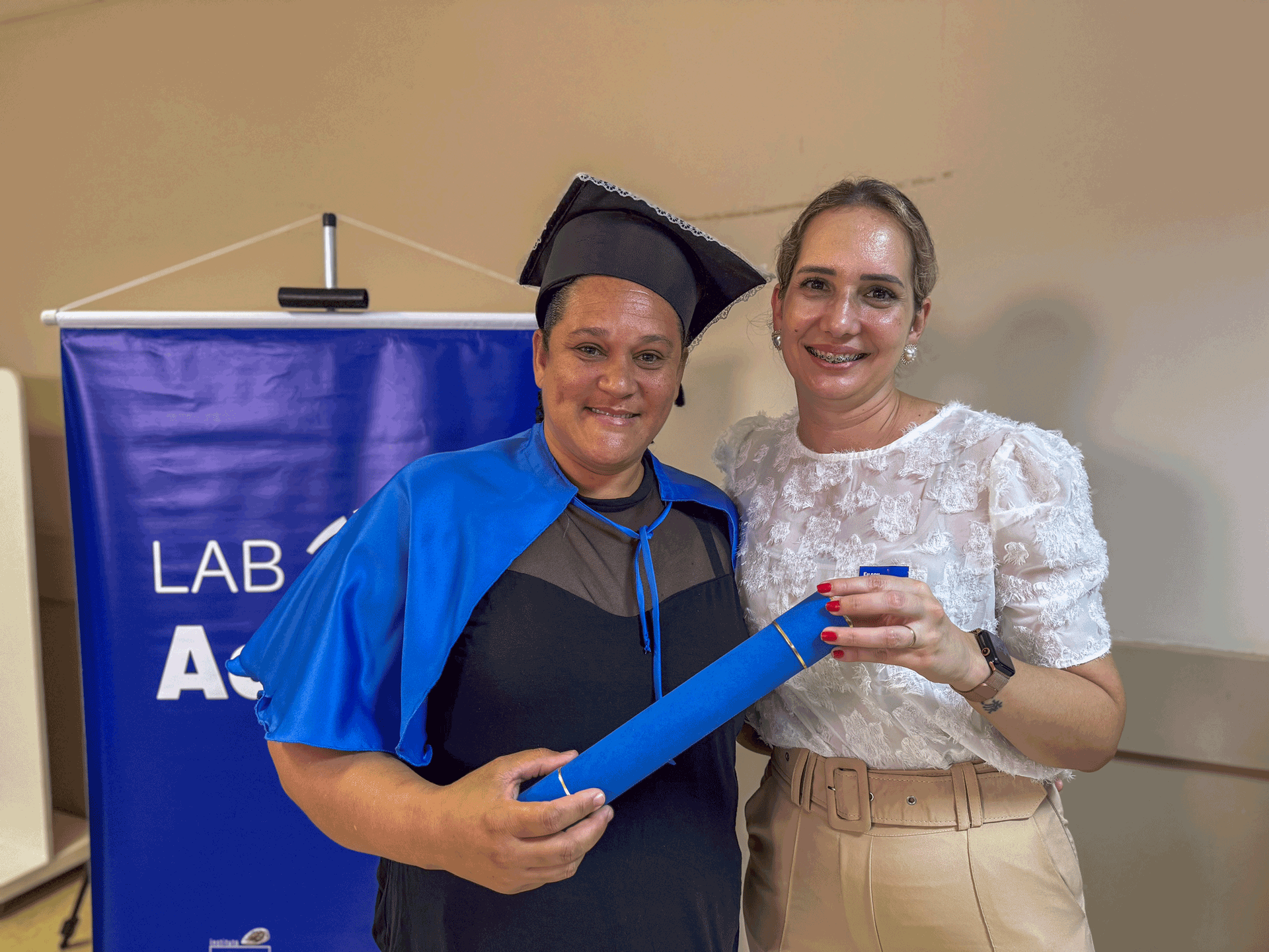 Duas mulheres sorrindo, uma delas com beca/capote de formatura, segurando diploma, ao lado de uma faixa.