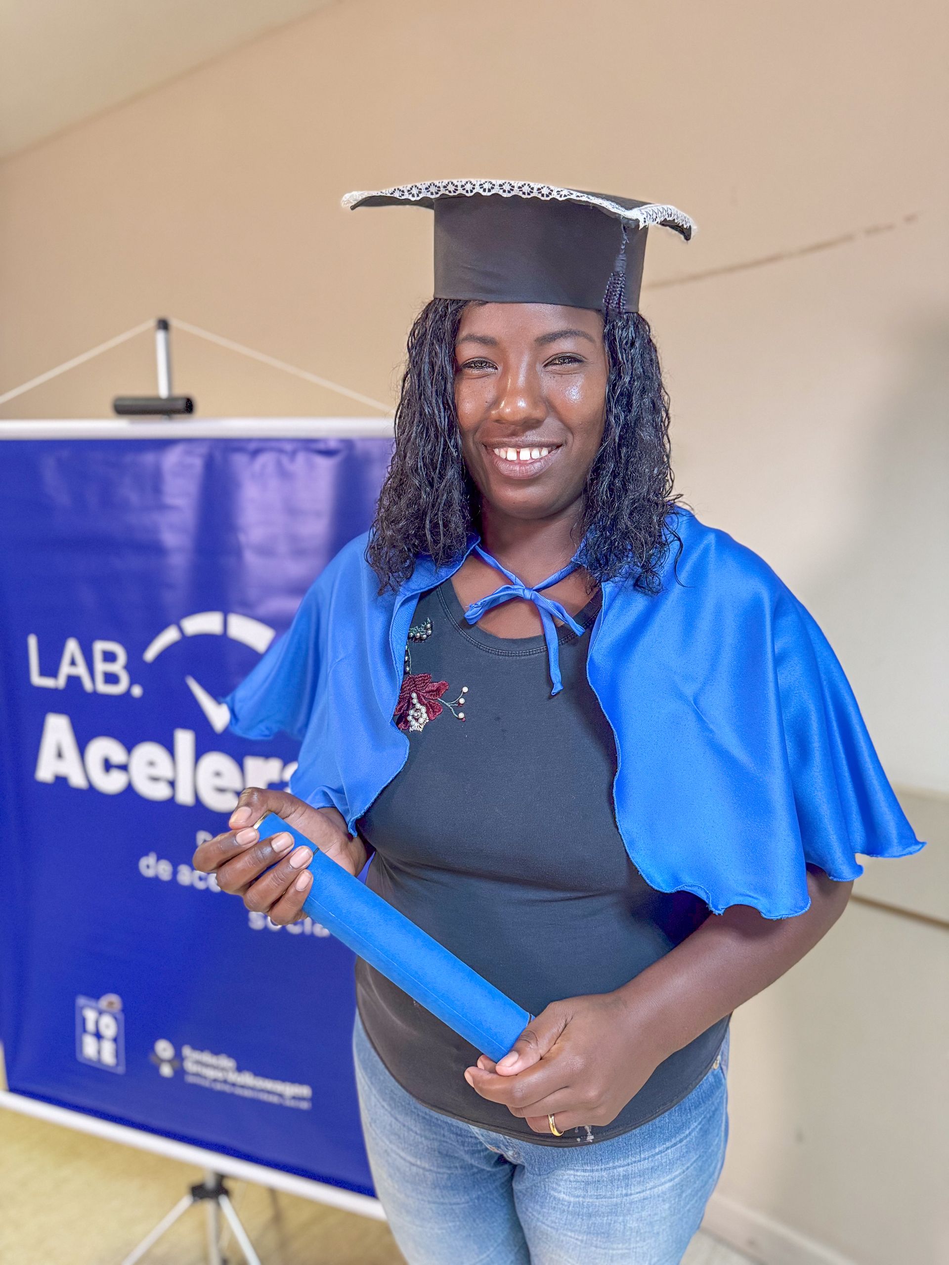 Mulher de beca e capelo de formatura, segurando um diploma, sorrindo em frente a uma faixa azul.
