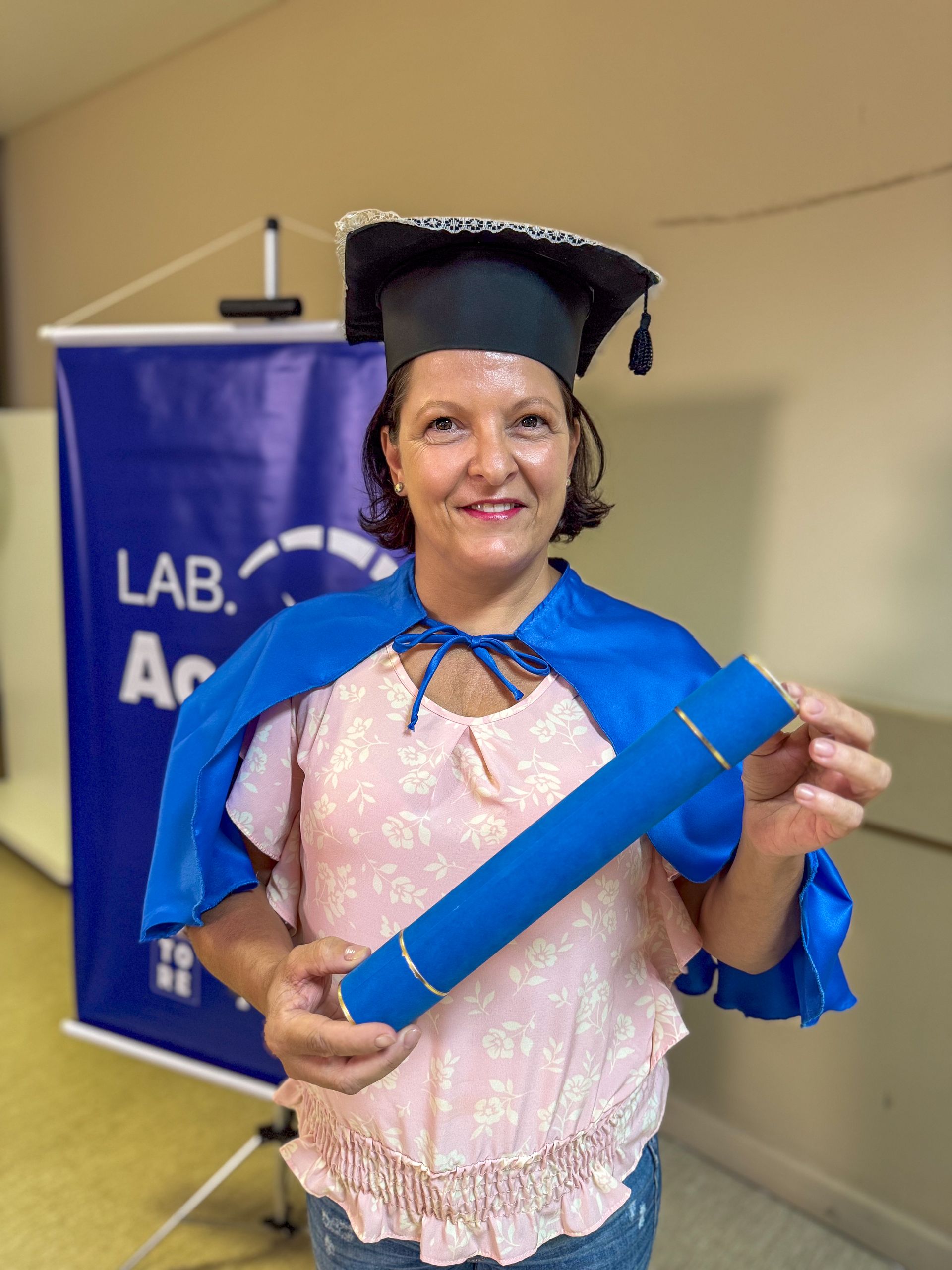 Mulher de chapéu e capa de formatura, segurando o diploma, sorrindo. Faixa azul em ambiente interno.