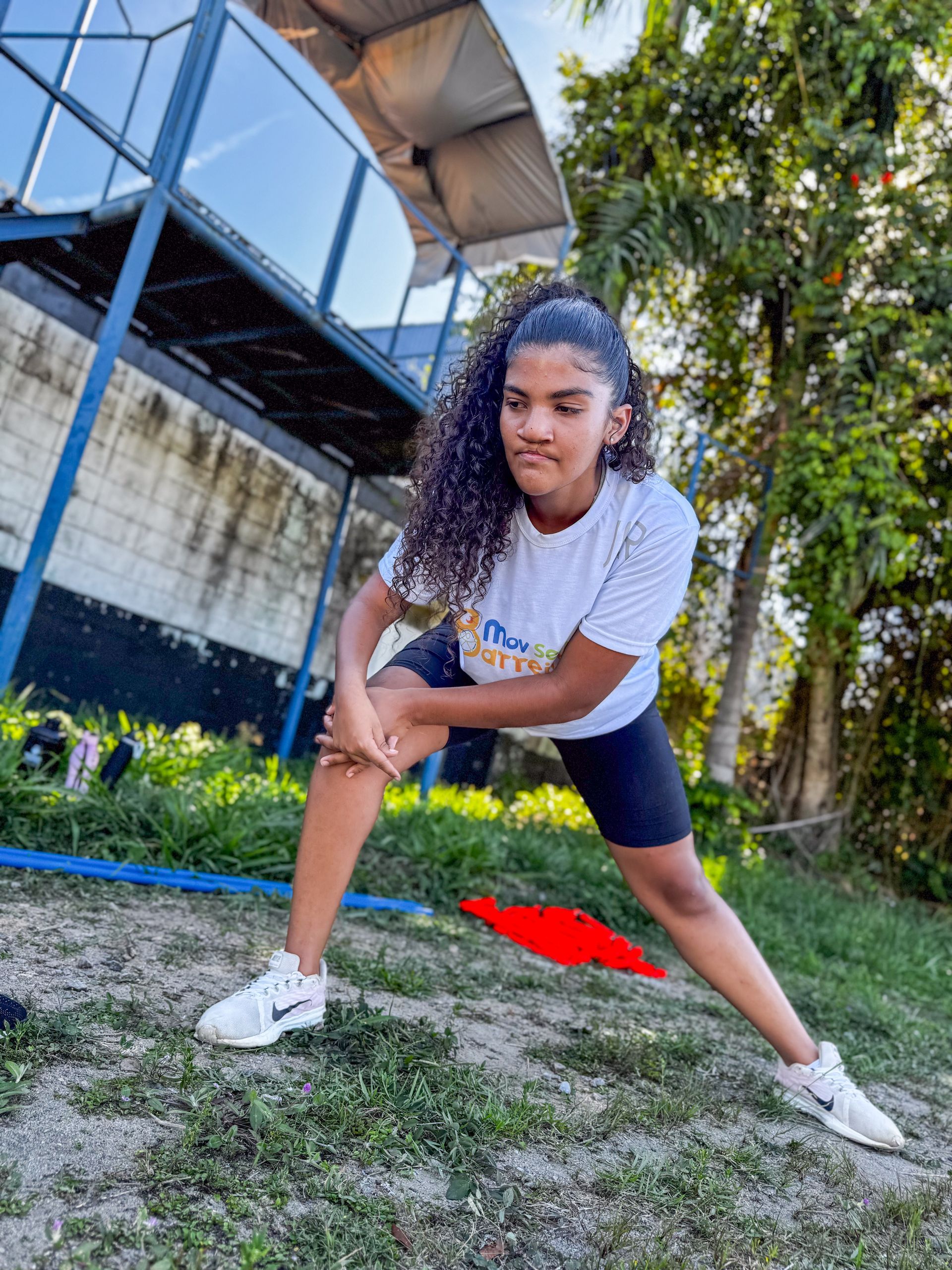 Uma jovem mulher com cabelos cacheados se esticando ao ar livre, pernas abertas, vestindo roupas esportivas.
