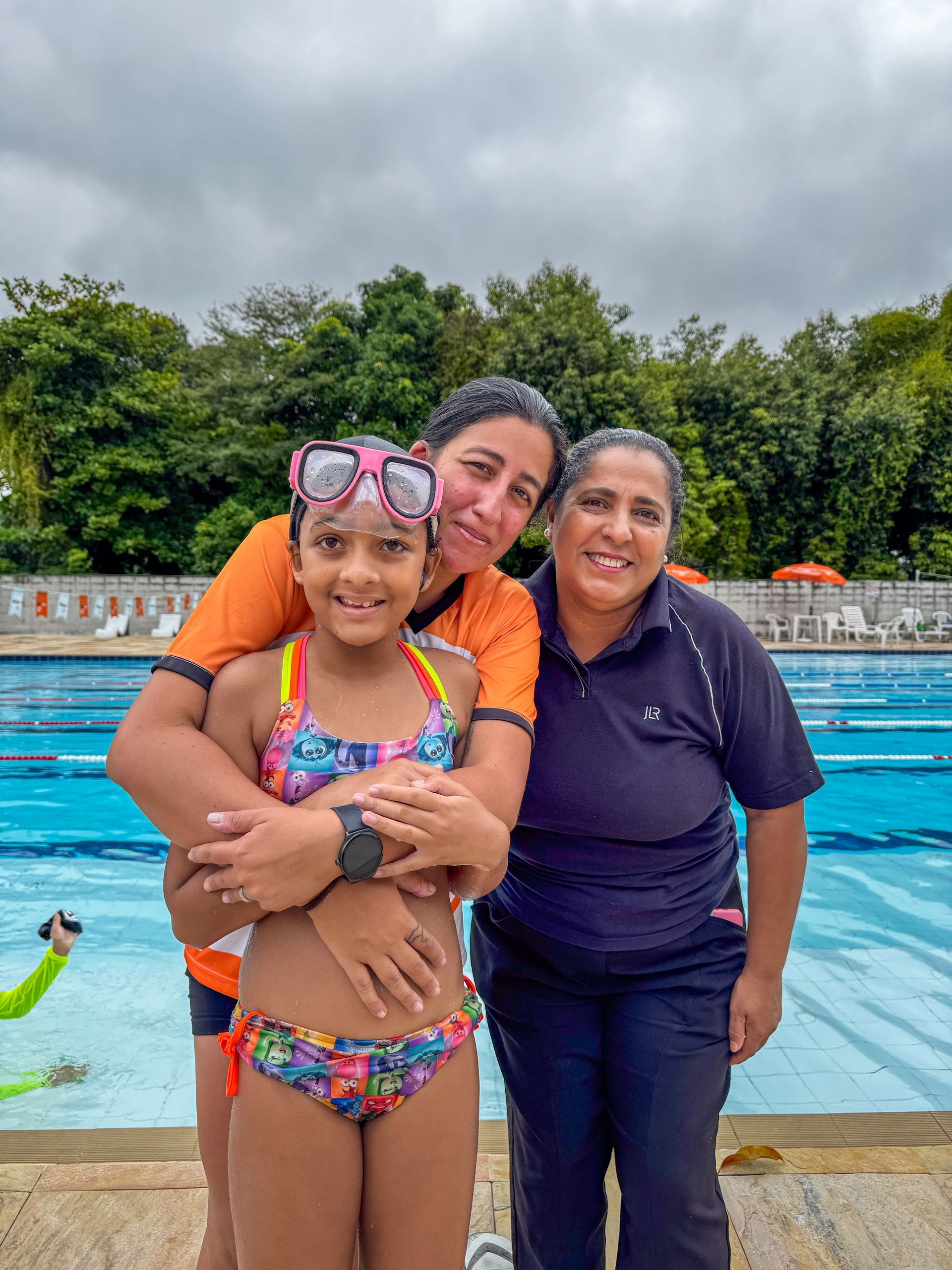 Uma garota de maiô é abraçada por duas mulheres perto de uma piscina em um dia nublado.