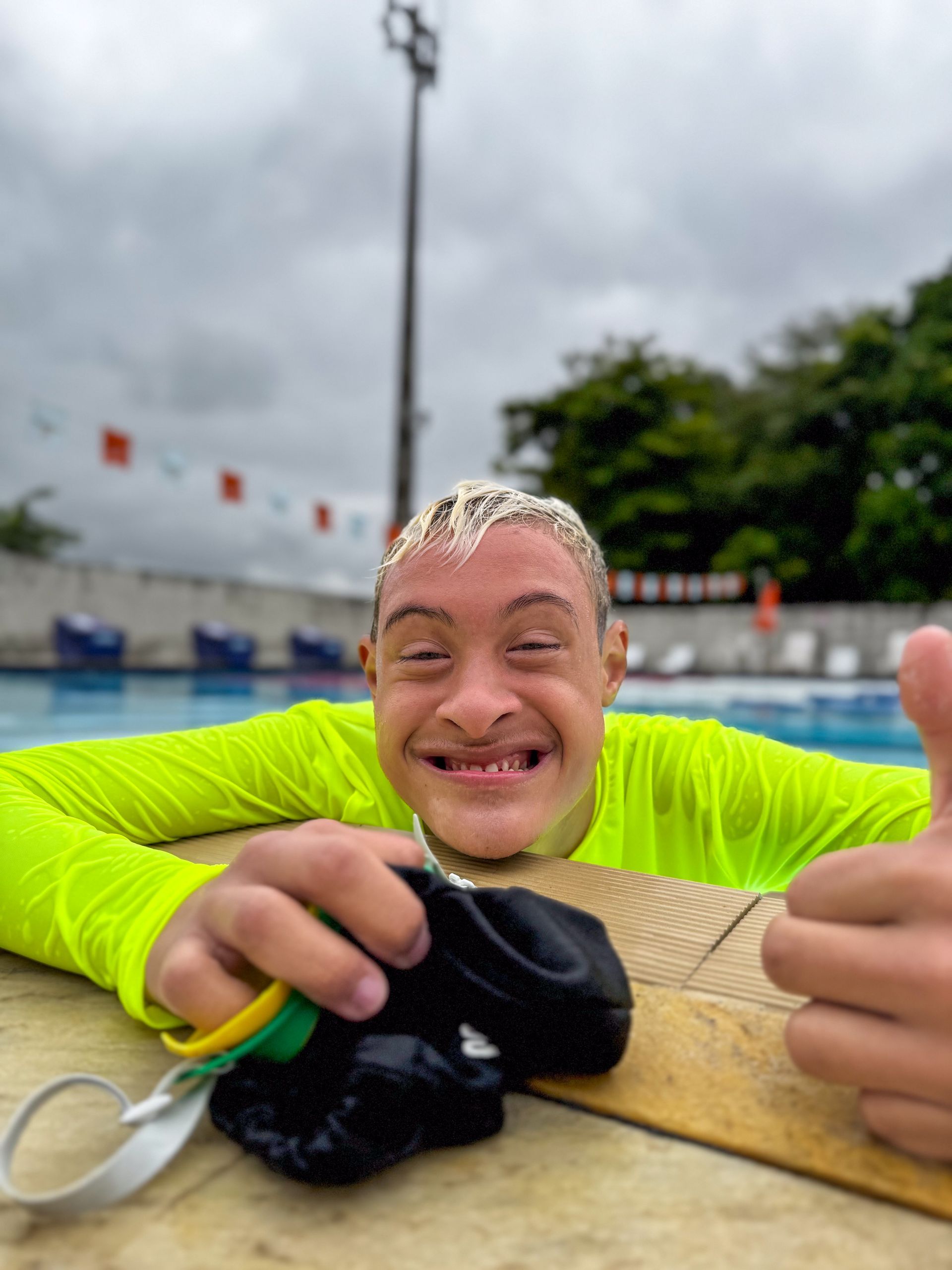 Homem com síndrome de Down, vestindo uma camisa amarela brilhante, sorri para a piscina e faz sinal de positivo.