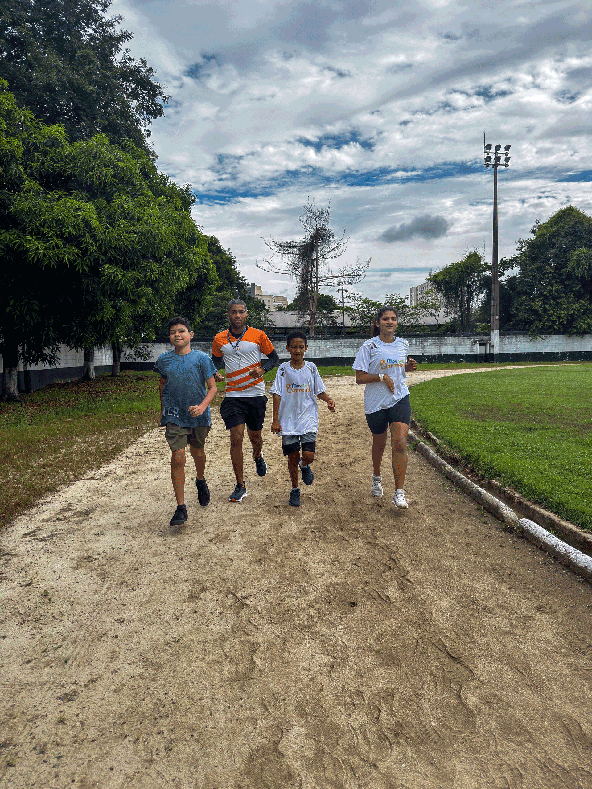 Quatro pessoas correndo em uma trilha de terra em um ambiente externo com árvores e um céu nublado.