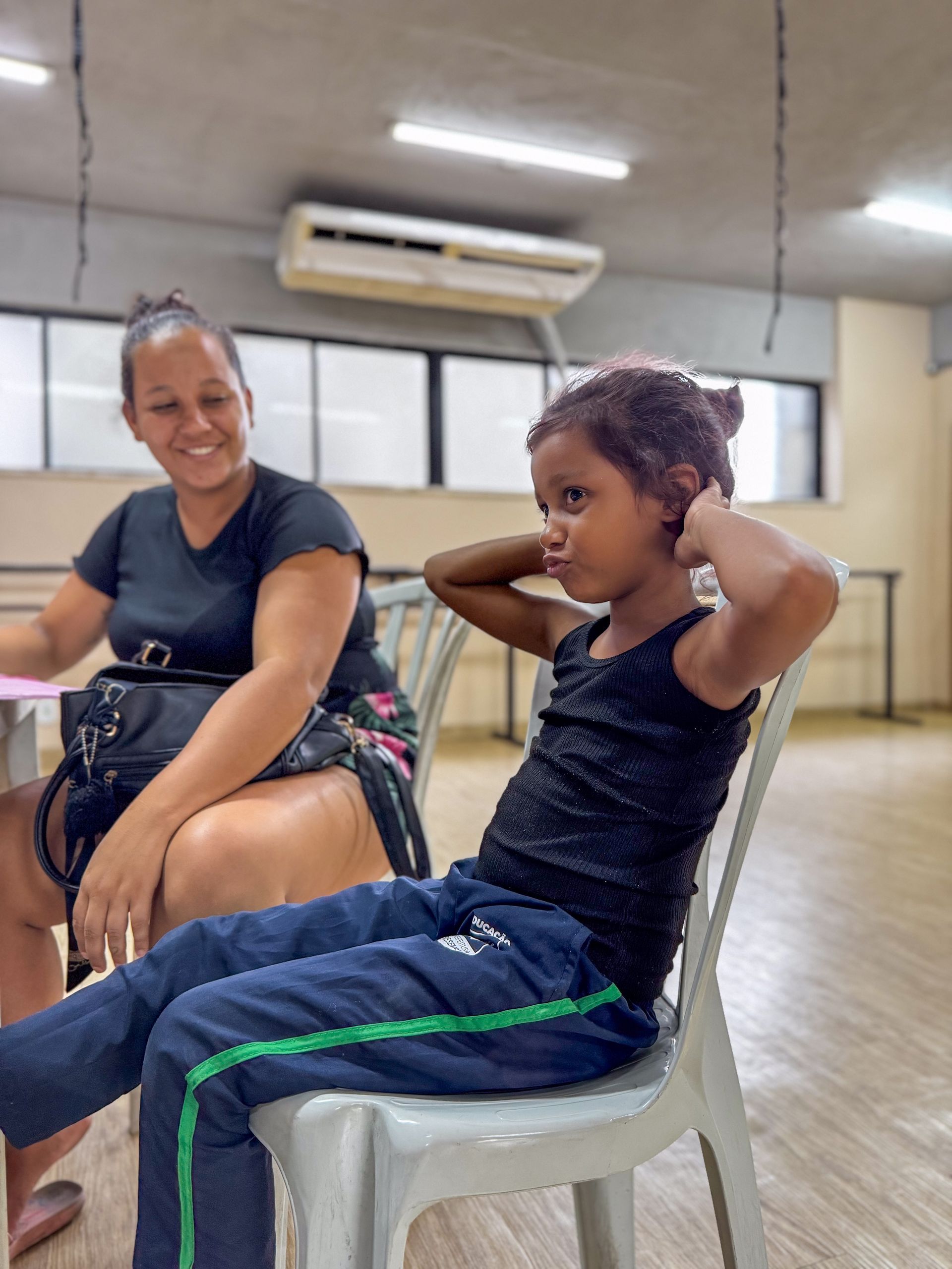 Uma mulher sorri enquanto uma criança está sentada em uma cadeira com as mãos atrás da cabeça dentro de casa.