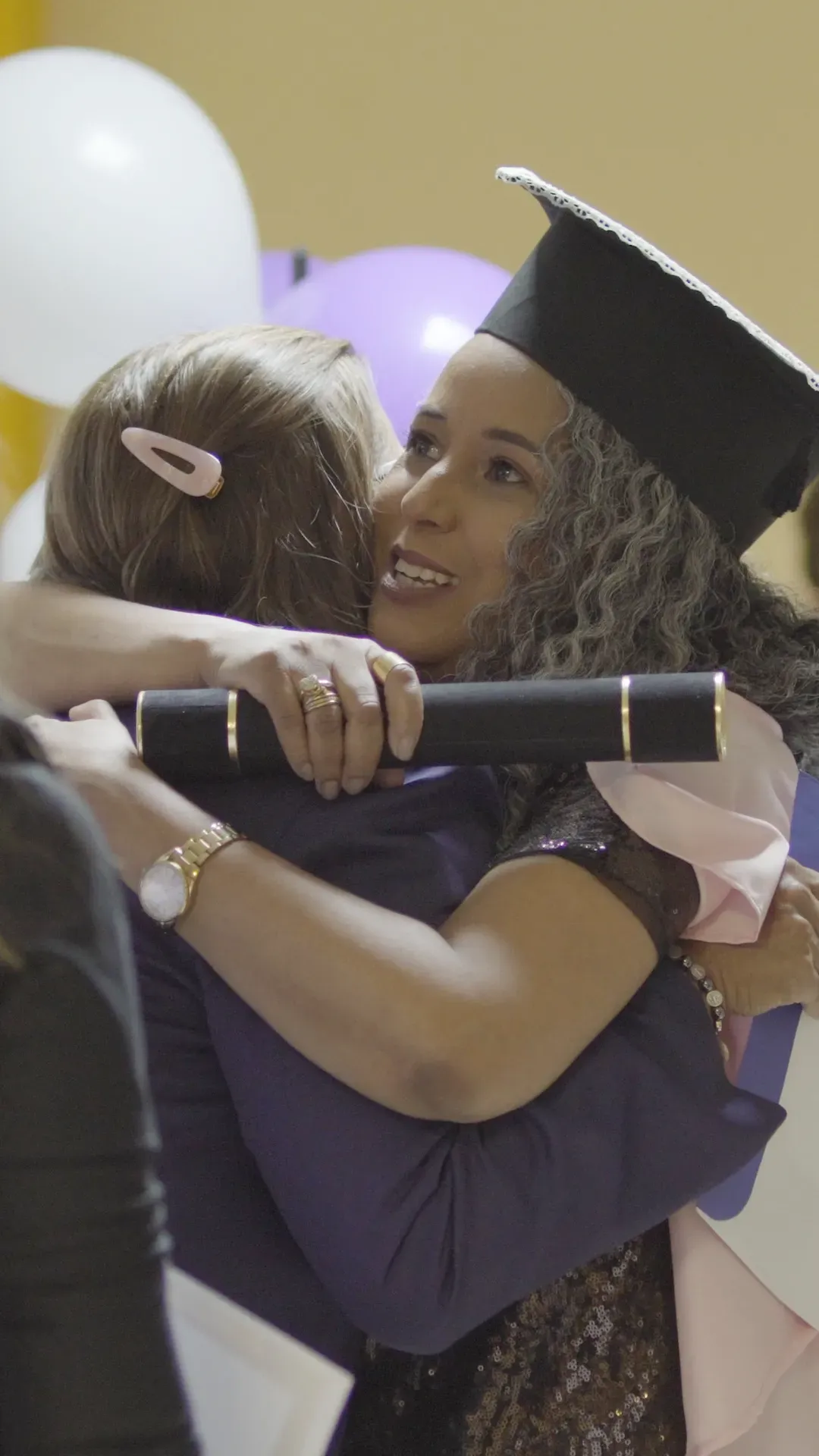 Mulher de boné abraça outra, que segura um diploma, sorrindo. Balões roxos e brancos e cenário.