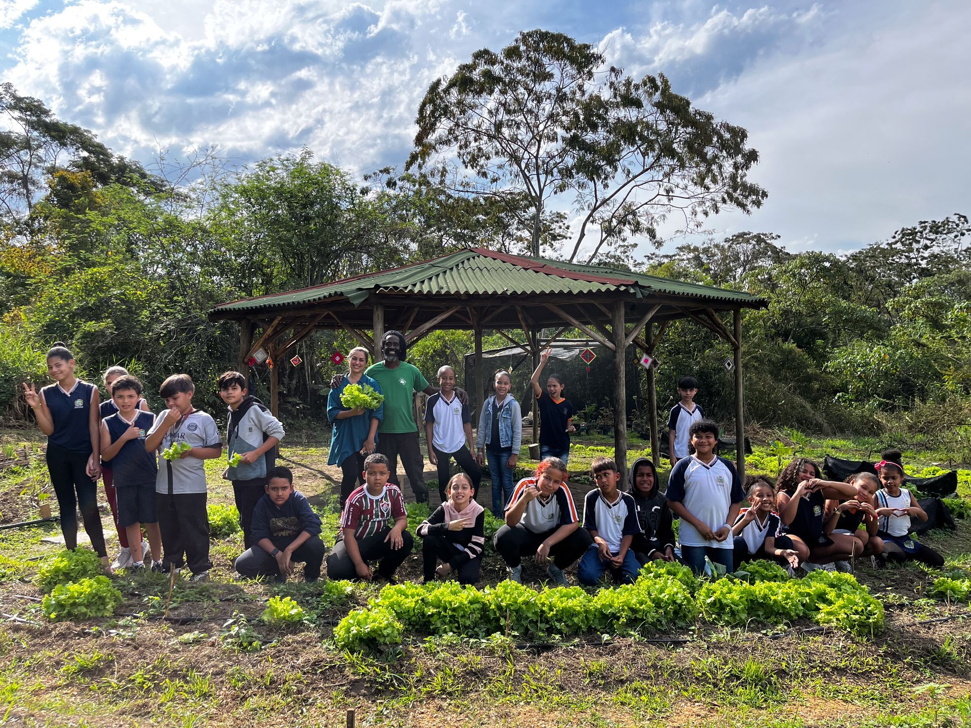 Grupo de crianças com uma professora sorrindo em um jardim. Plantas verdes, terra marrom, um gazebo e árvores ao fundo.