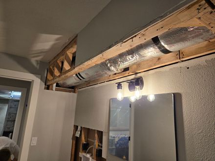 An exposed ceiling soffit reveals ductwork and framing above a bathroom vanity with a mirror and three-light fixture.