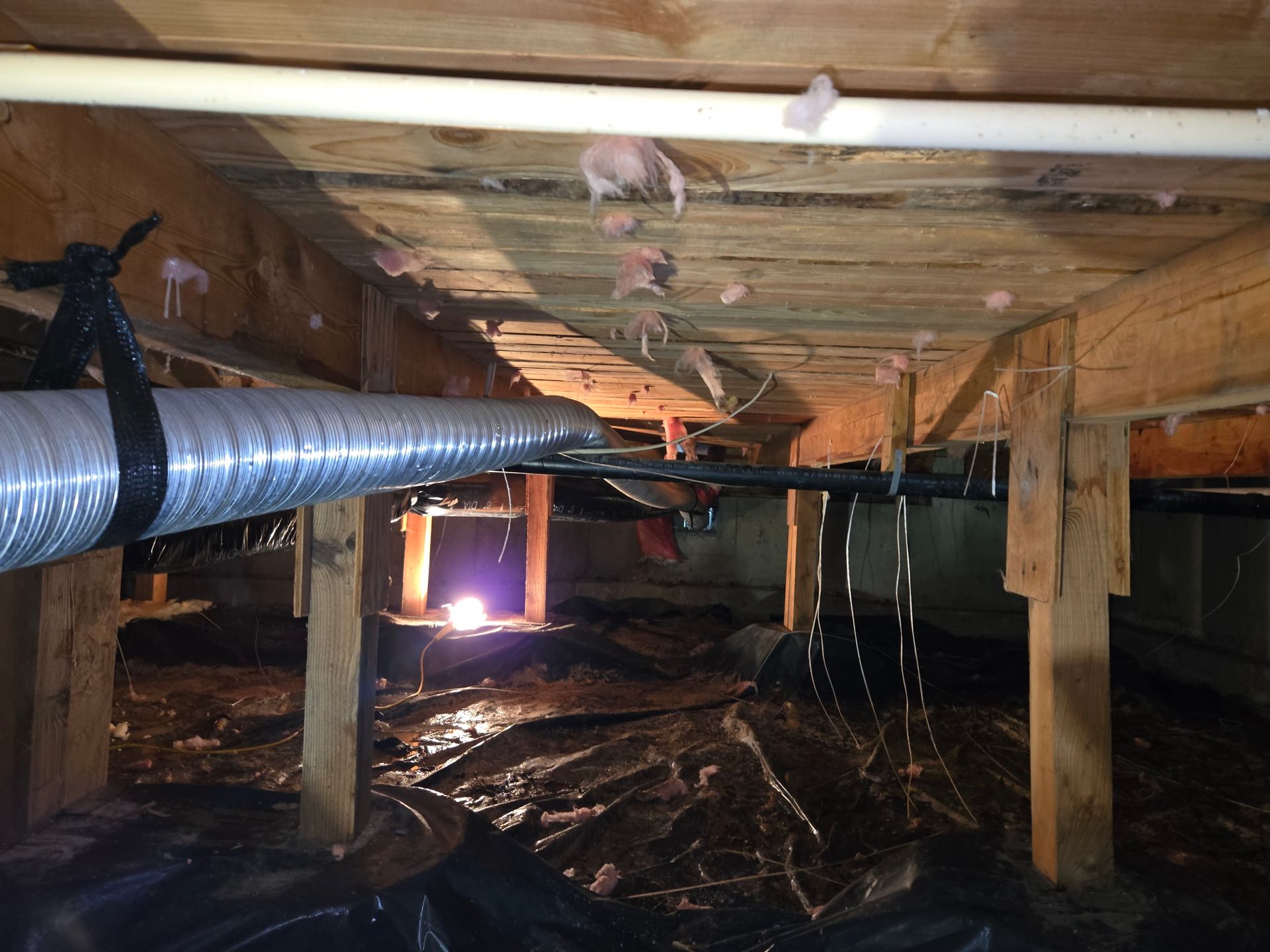 A low-angle view of a damp, unfinished crawl space with exposed wooden beams, insulation, plastic sheeting, and ducting.
