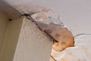 Water-damaged ceiling plaster peeling away to reveal rusted metal reinforcing bars near a pillar.