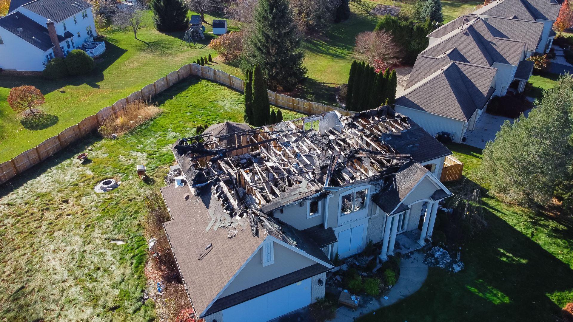 An aerial view of a residential house with a severely fire-damaged roof, exposing the charred wooden frame.