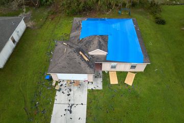 Aerial view of a residential house with roof damage covered by a large blue tarp, and debris scattered on the driveway.