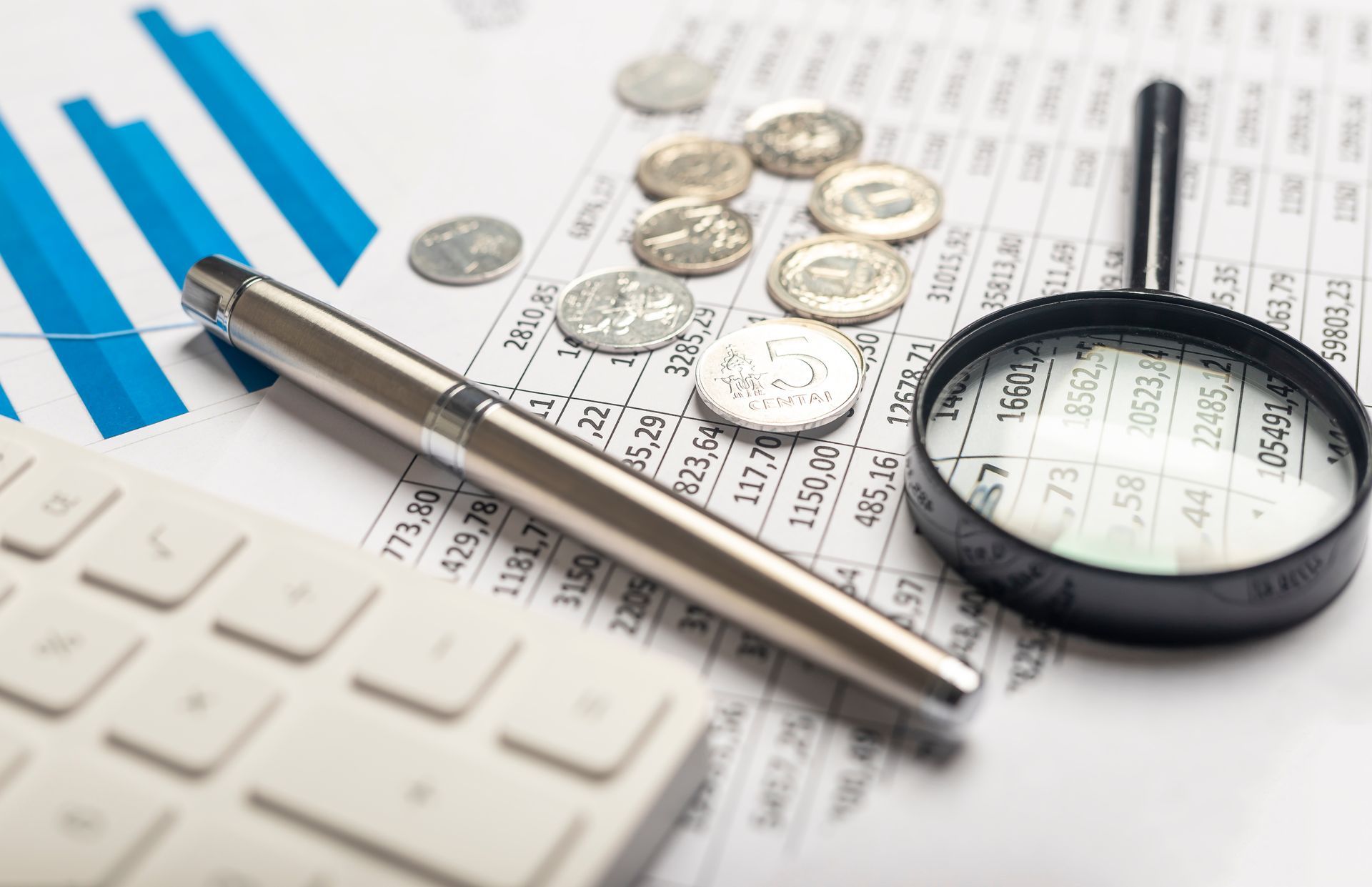 Business calculator, pen, coins, and magnifying glass on financial charts and keyboard.