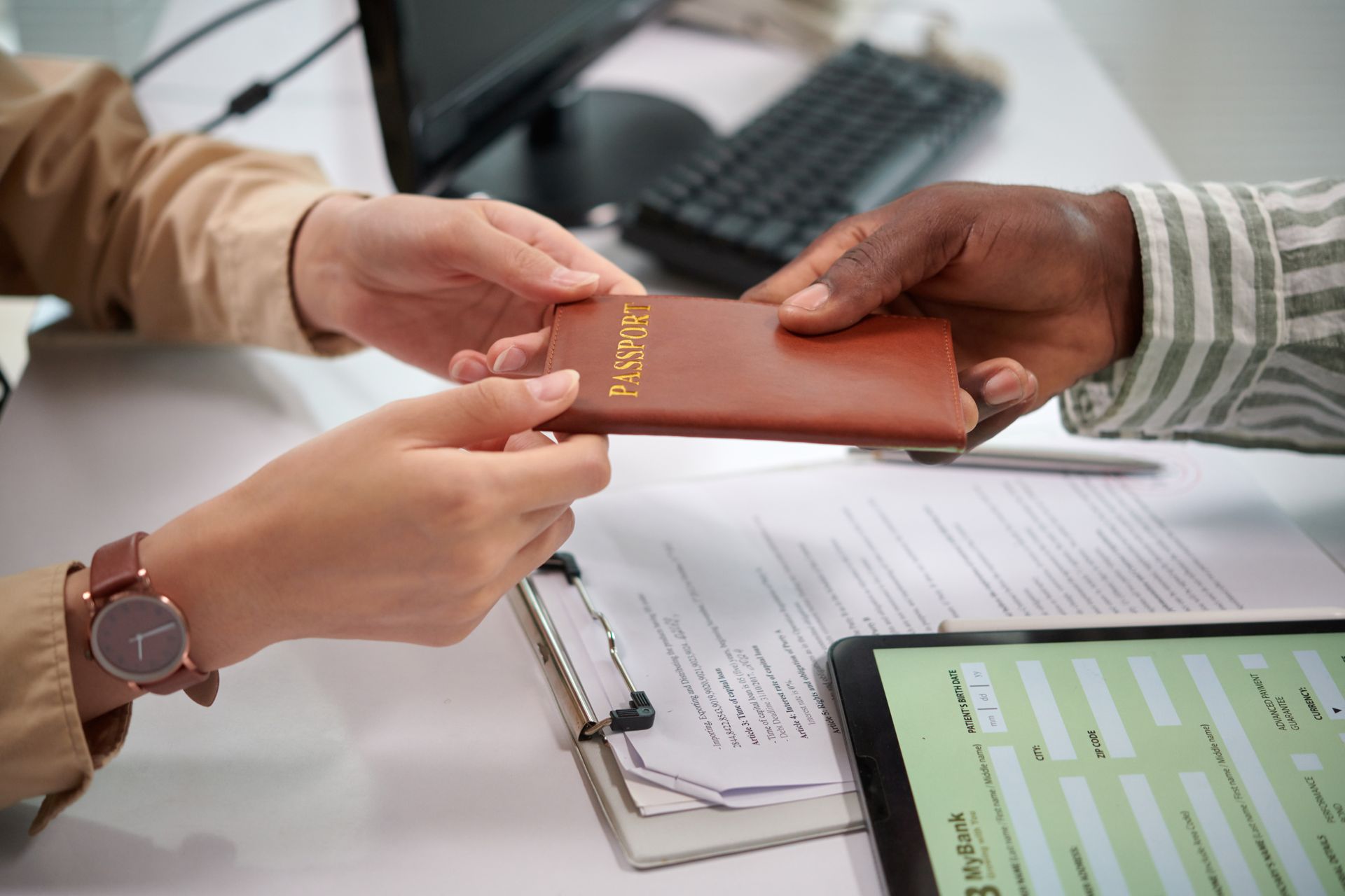 Hands exchanging a brown passport over a desk with forms and a laptop, suggesting travel or immigration processing