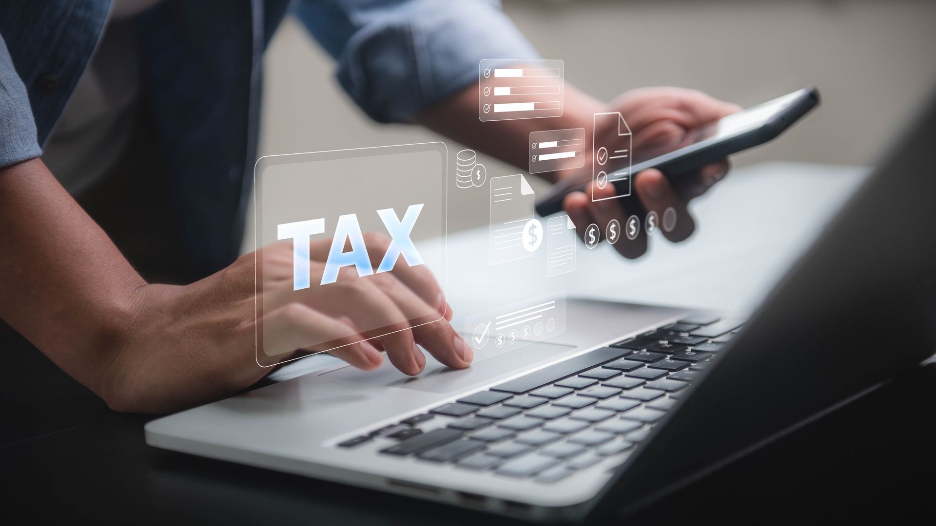 Hands using laptop and smartphone with digital tax icons floating above the keyboard