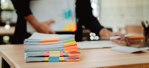Stack of colorful paper files on a desk with blurred people working in the background