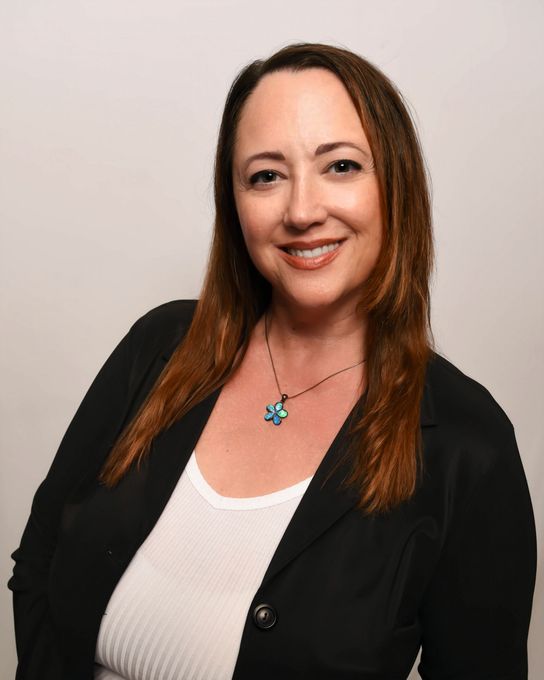 Woman with long brown hair, wearing a black blazer over a white shirt, smiling, and wearing a necklace.