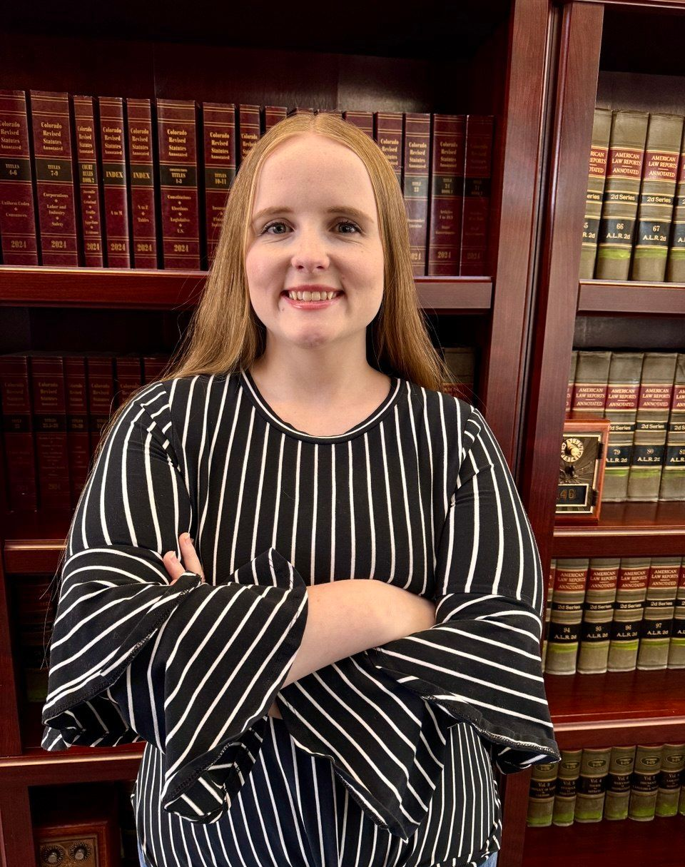 A person with long reddish hair smiles with arms crossed, wearing a black-and-white striped blouse in a library.