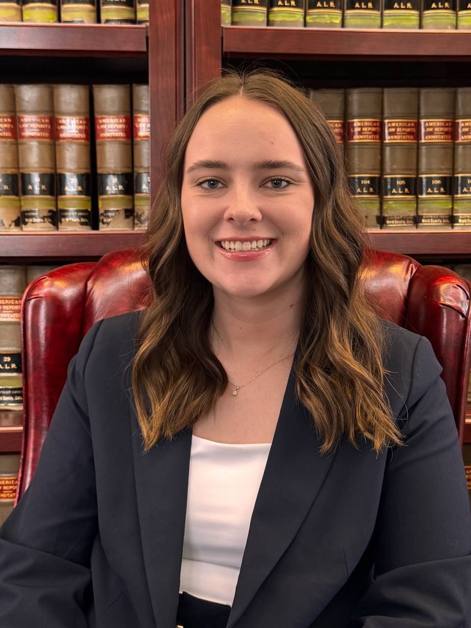 Woman in a navy blazer seated in a leather chair, smiling in front of law books on wooden shelves.