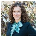 Woman with curly brown hair, wearing a dark blazer and teal scarf, smiles outside.