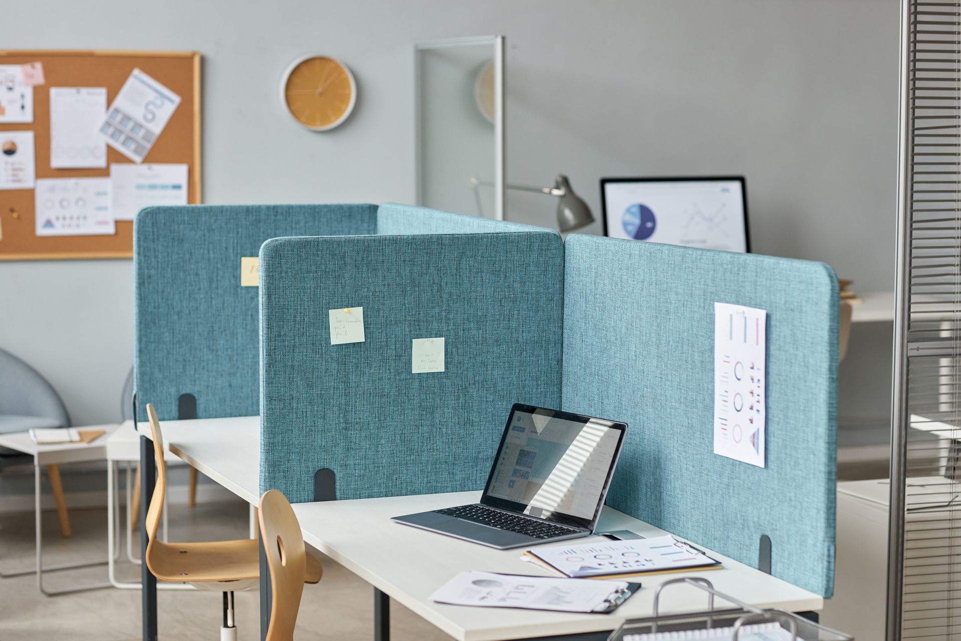 Office desk with blue fabric partitions, a laptop, papers, and a wall corkboard in a bright workspace