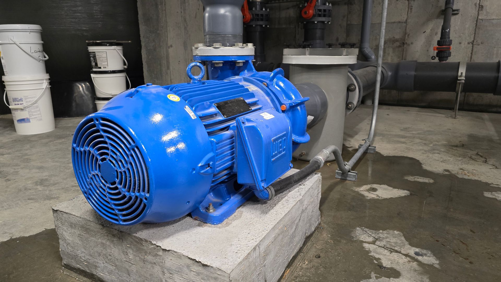 Moteur électrique industriel bleu sur un bloc de béton, relié à des tuyaux. Environnement d'une buanderie.